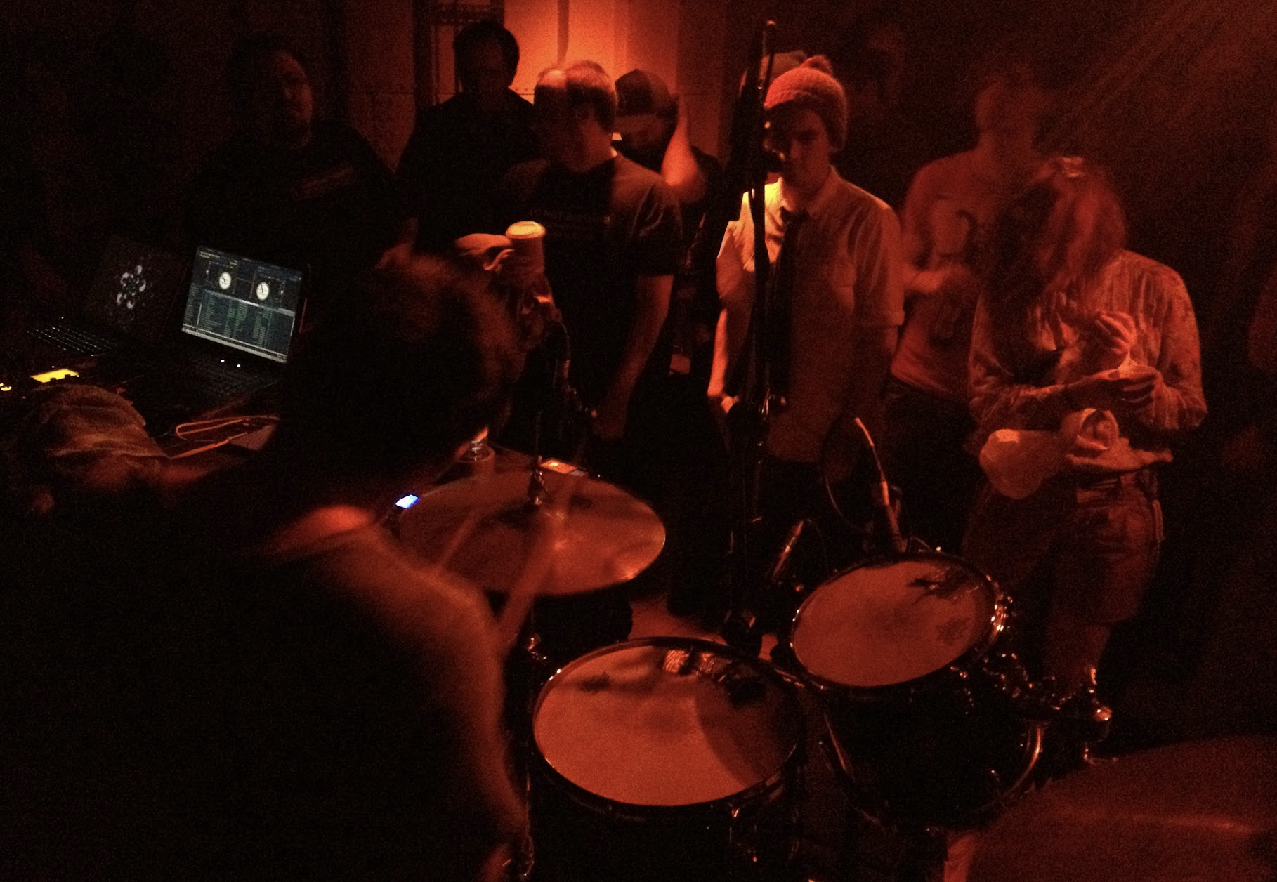 A dimly lit scene with a group of people in a recording studio or music venue, some standing and others seated around a drum set, with a person playing drums and a laptop with music software nearby.