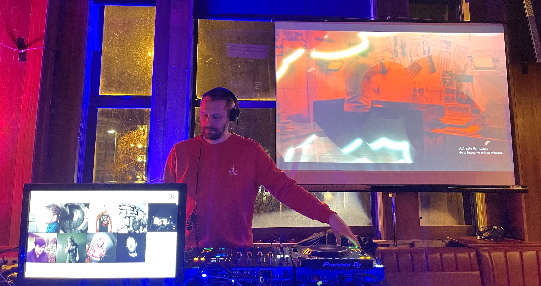 A DJ with headphones standing at a turntable in a dimly lit room with colored neon lights; a large screen behind him displays abstract visuals and a smaller screen in front shows a grid of photos of people.