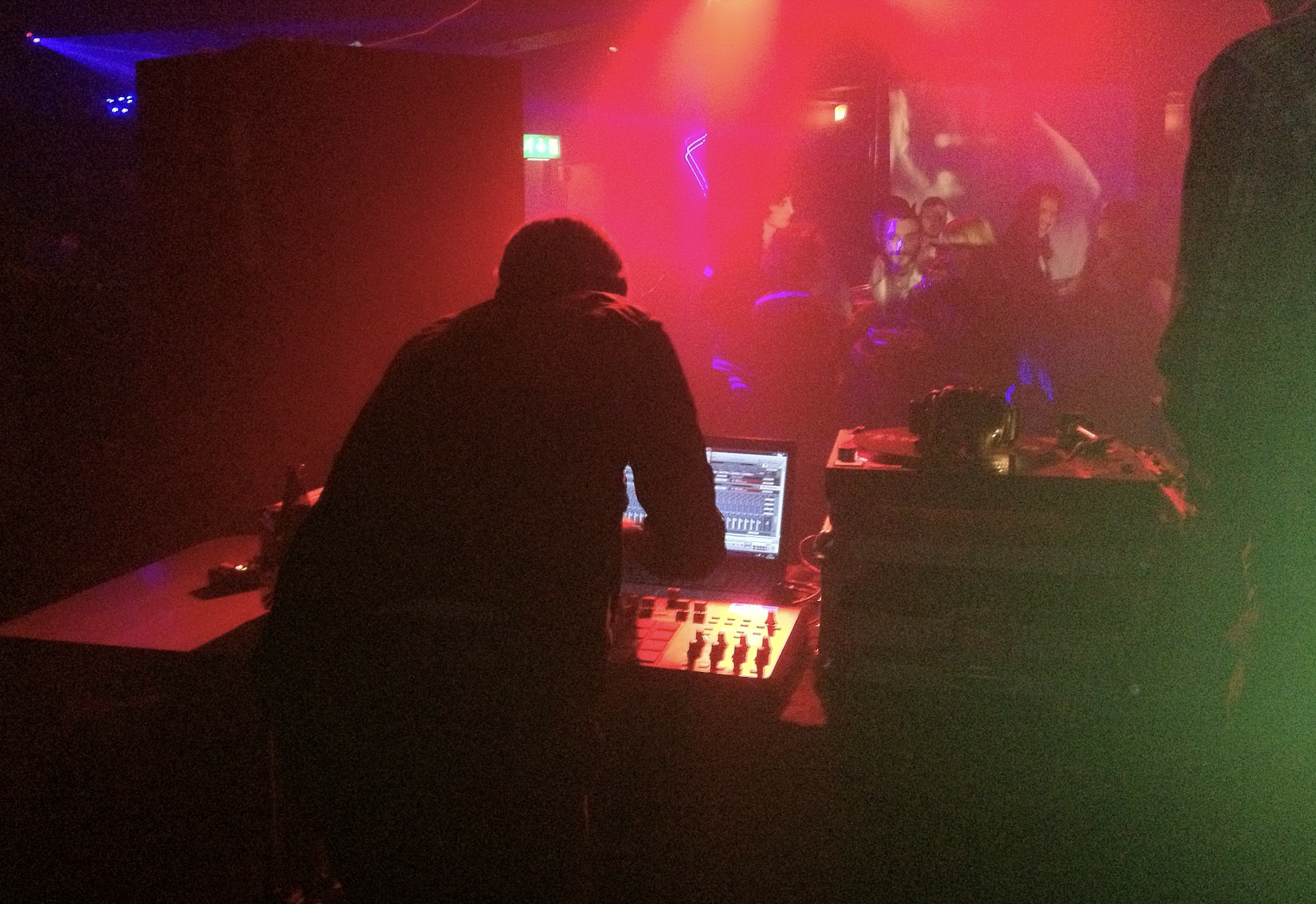 A DJ performing in a dimly lit club with red and purple neon lights, people dancing in the background.