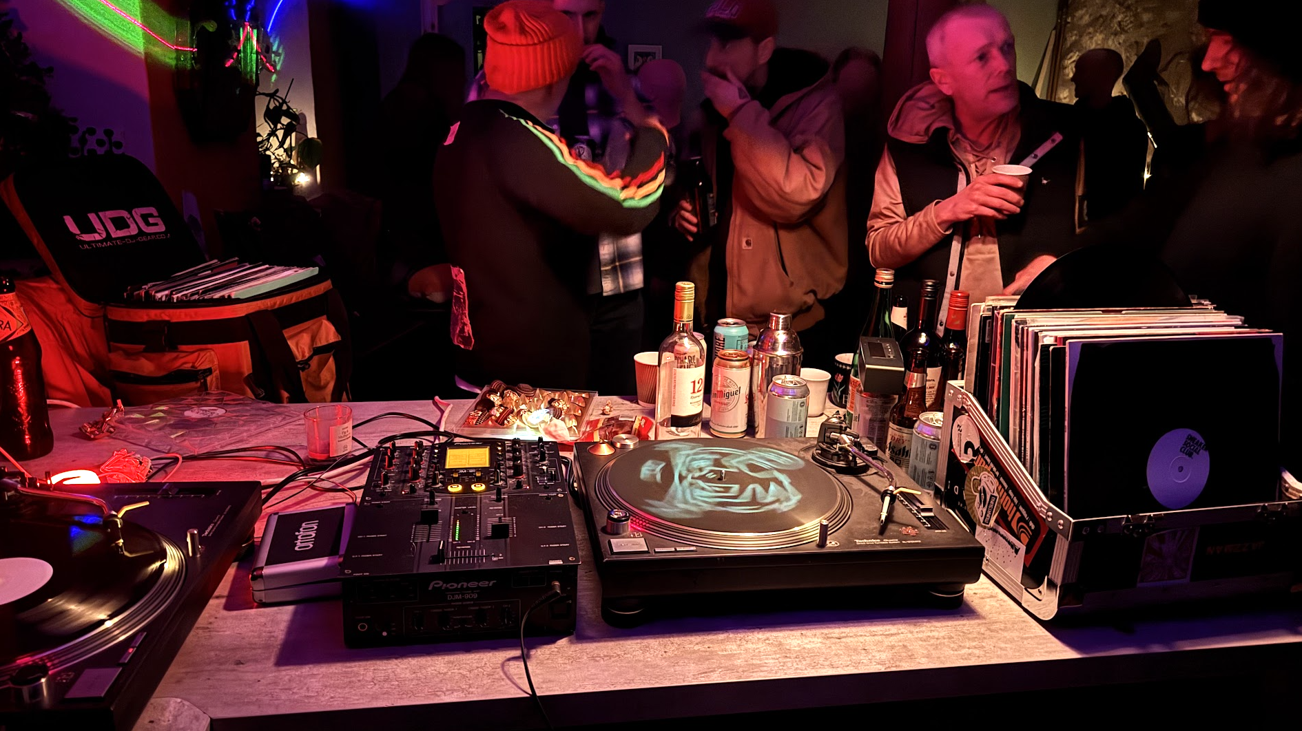 DJ setup with vinyl records, turntable with a face image, mixer, and drinks, with people socializing in the background at a party.