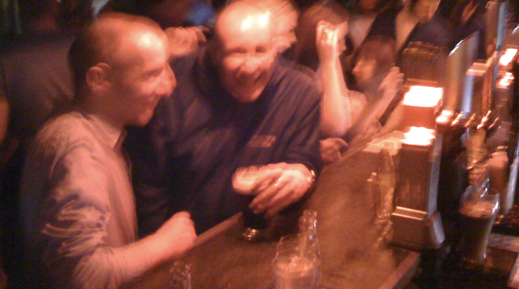Two men sitting at a bar, laughing and enjoying drinks in a lively setting.