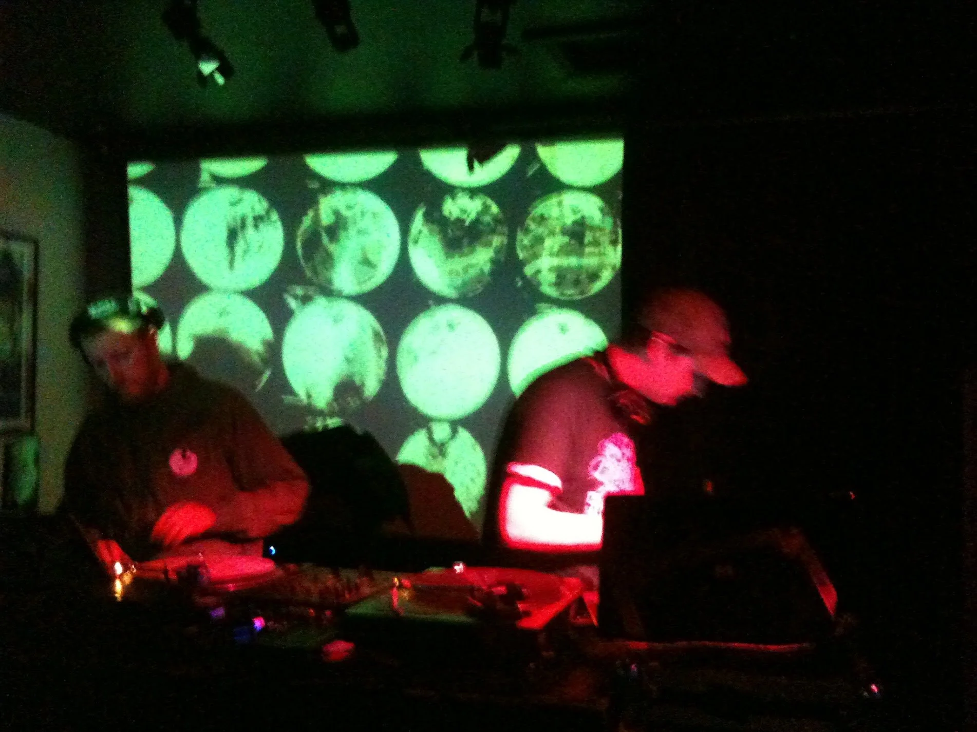 Two DJs wearing headphones and baseball caps perform in a dark room with a green circular patterned projection behind them.