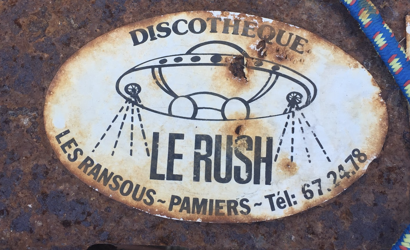 Rusty oval sign with a UFO illustration and French text for a discotheque named 'Le Rush' in Pamiers, France, listing a phone number.