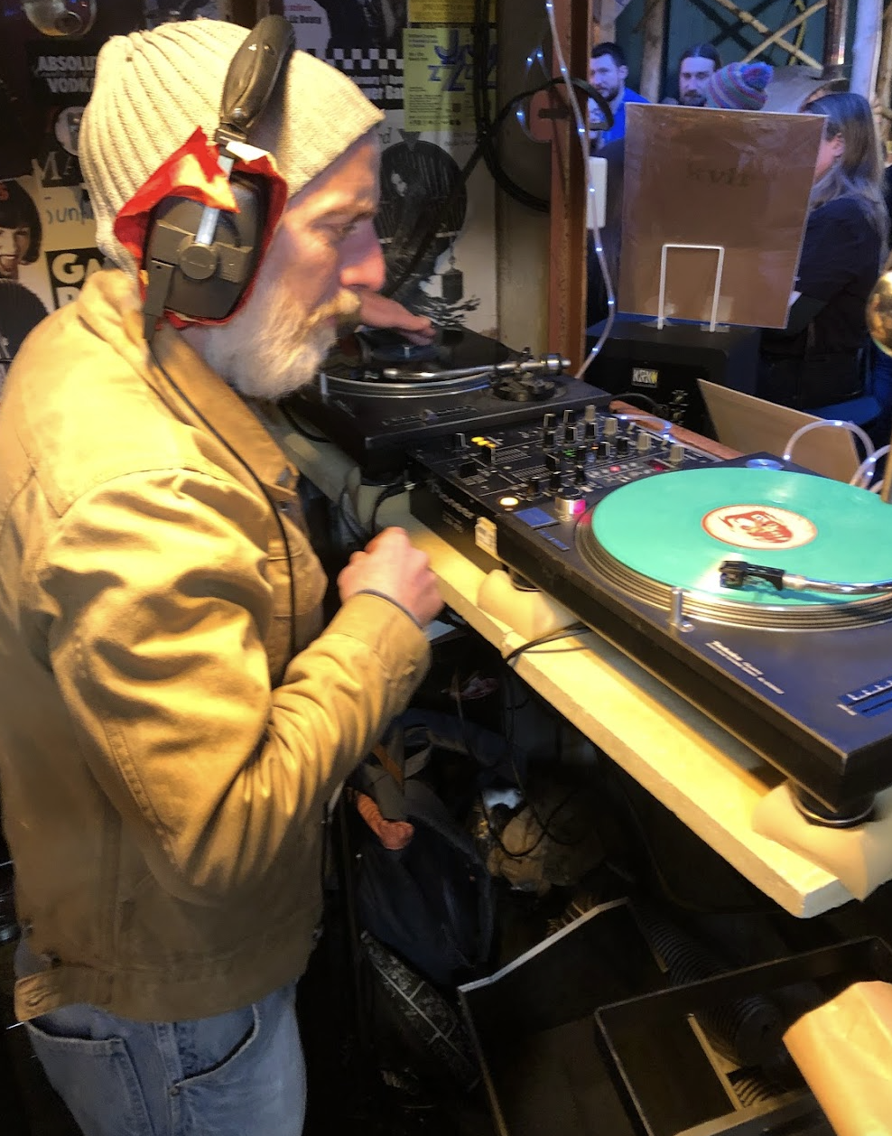 A man with a beard wearing a gray beanie, yellow jacket, and headphones, standing at a DJ turntable setup in a lively indoor setting with posters on the wall and people socializing in the background.