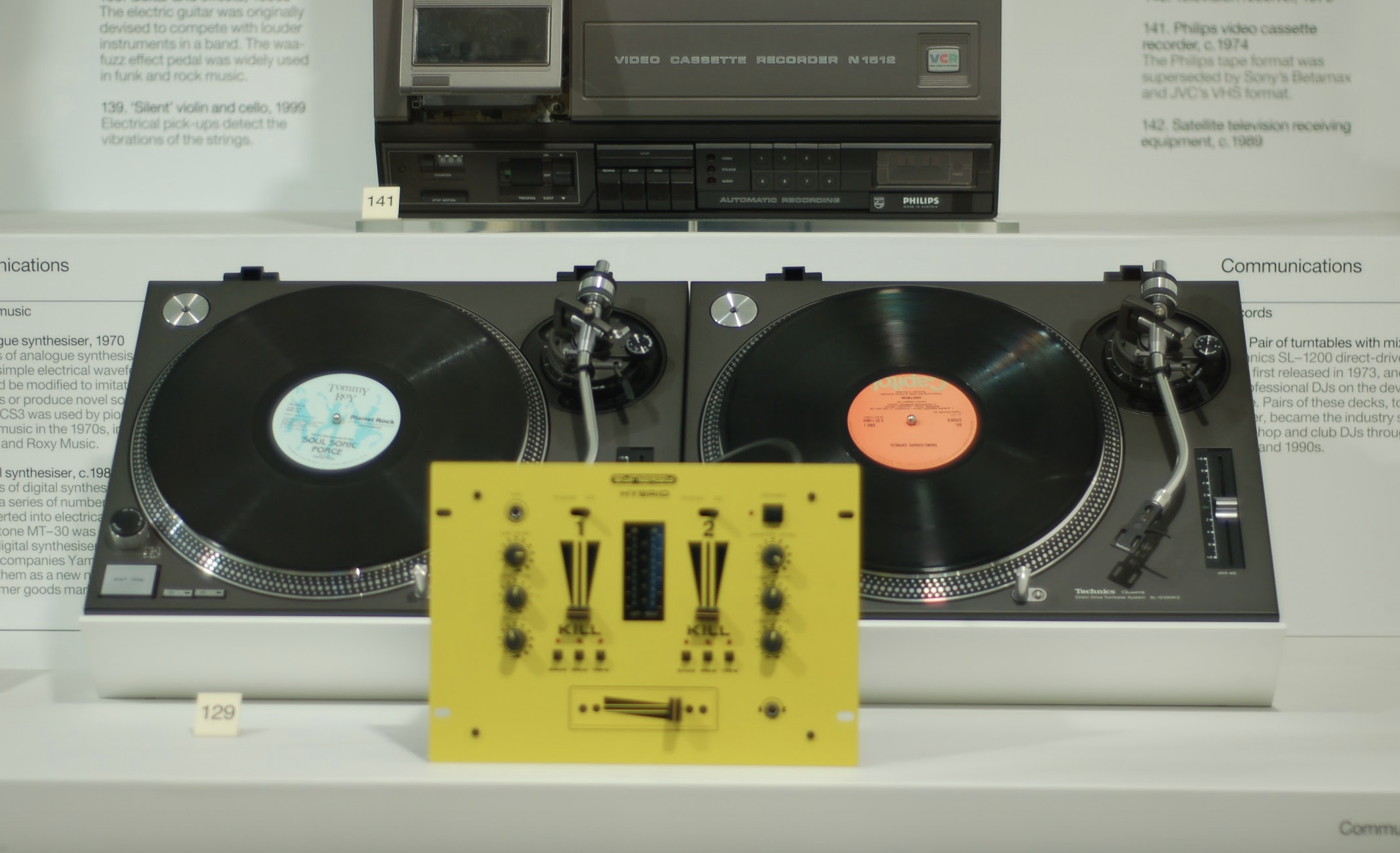 A turntable with two black vinyl records, set on a white surface, with a yellow DJ mixer in front. In the background, a vintage video cassette recorder and a television are displayed behind a glass case.