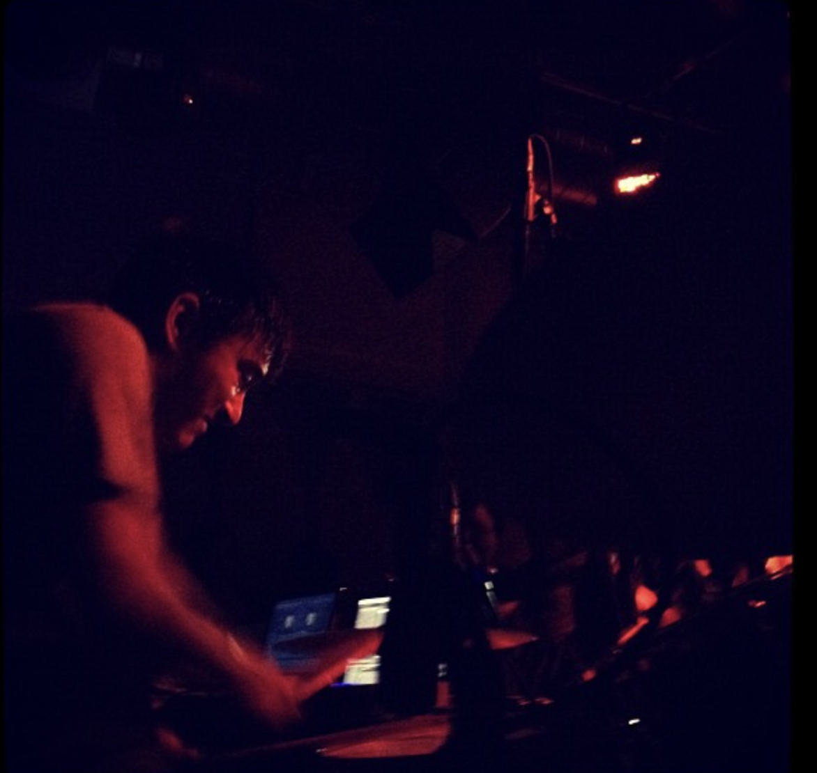 A DJ performing in a dark club with red lighting, looking down at equipment with a laptop in front of him.