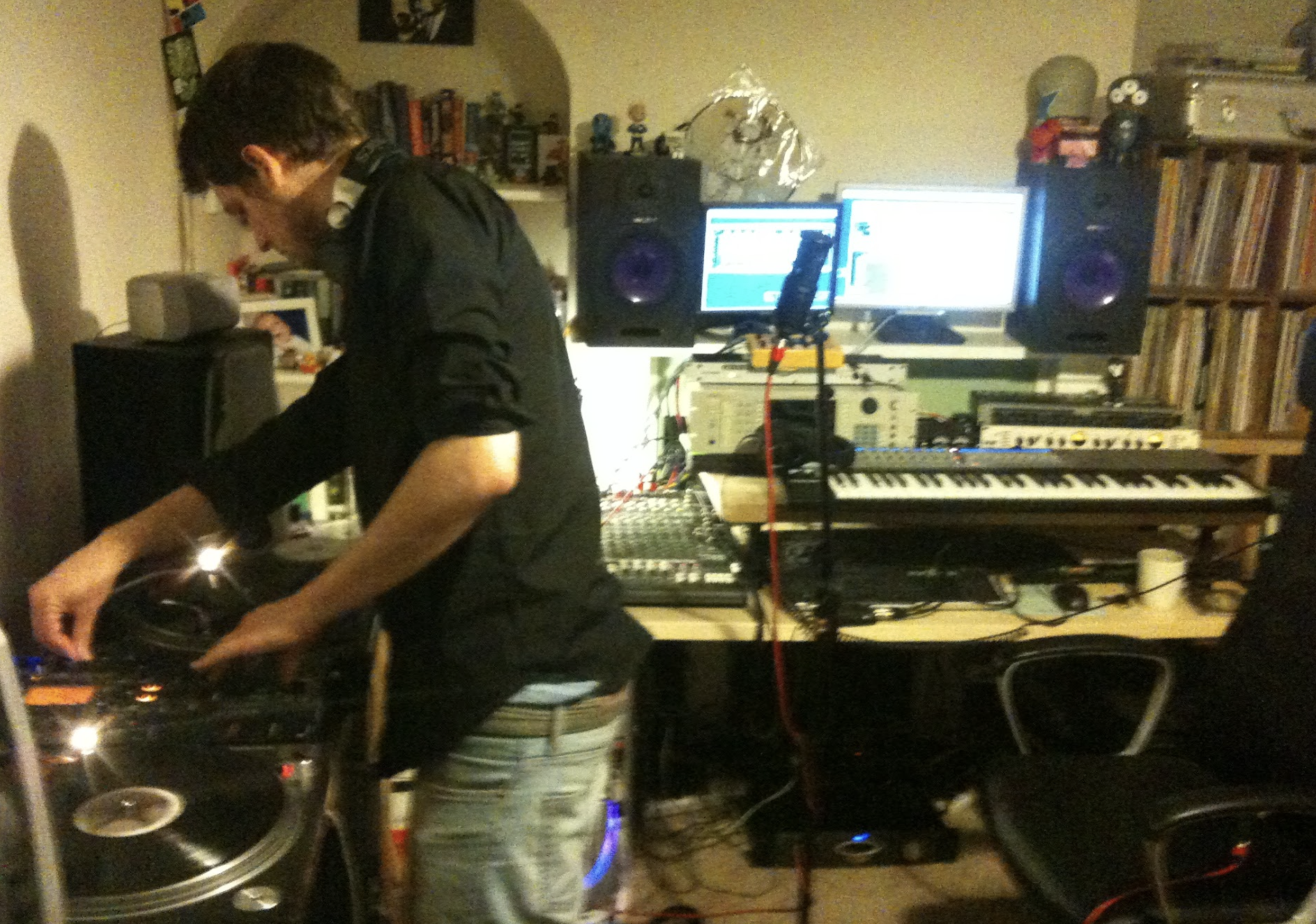 A person working in a home music studio with DJ equipment, turntables, a keyboard, speakers, computer monitors, and shelves filled with books and collectibles.