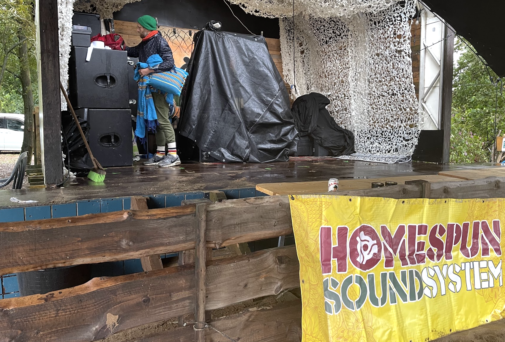 A person standing on a wooden outdoor stage with music equipment, covered in a black tarp, and a large yellow banner with the words 'HOMESPUN SOUNDSYSTEM'. The person is wearing a colorful jacket, green pants, and colorful socks, and is near the spea