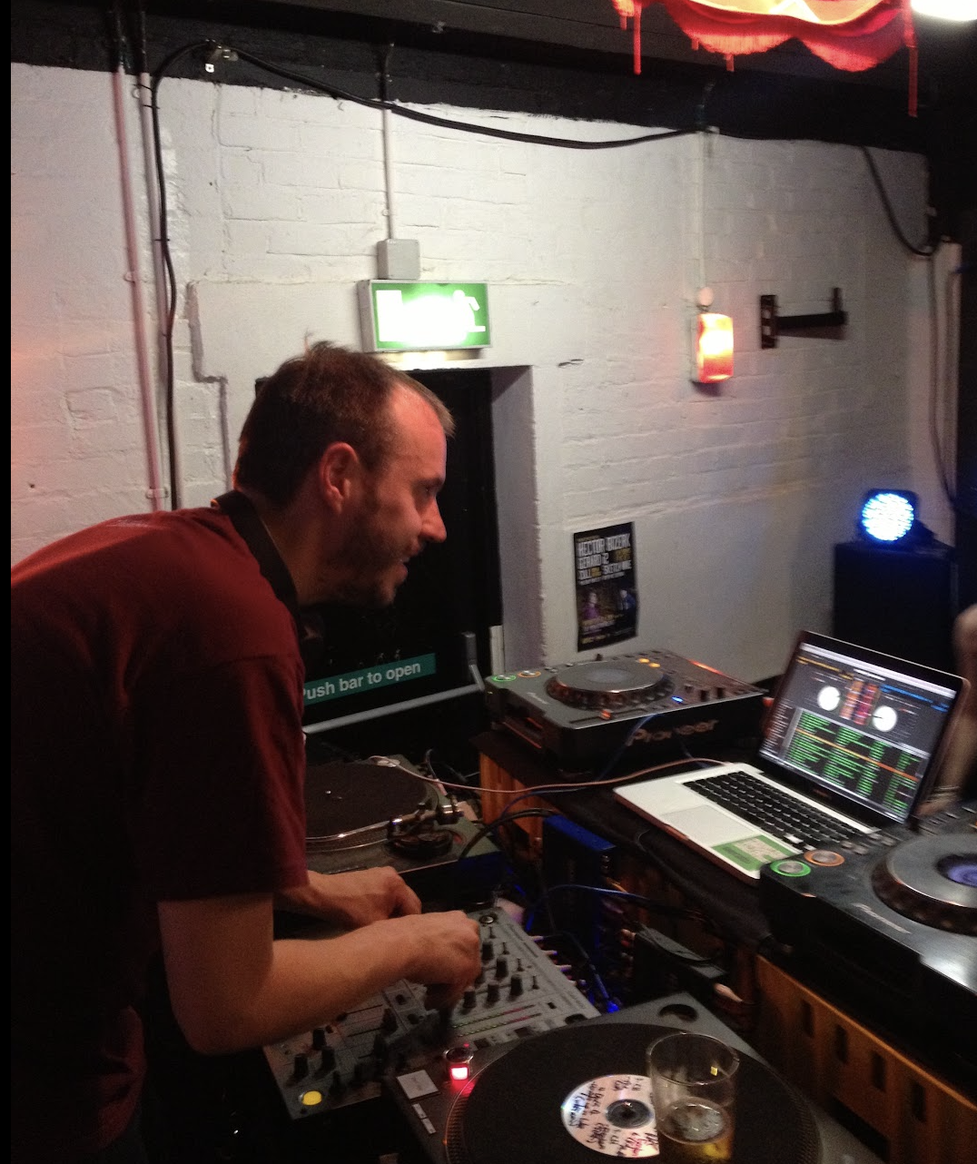 A DJ with short hair and a beard is performing in a dimly lit venue, using a DJ mixer connected to a laptop with music software on the screen. There is a glass of beer on a turntable in front of him, and the background includes a white brick wall, a