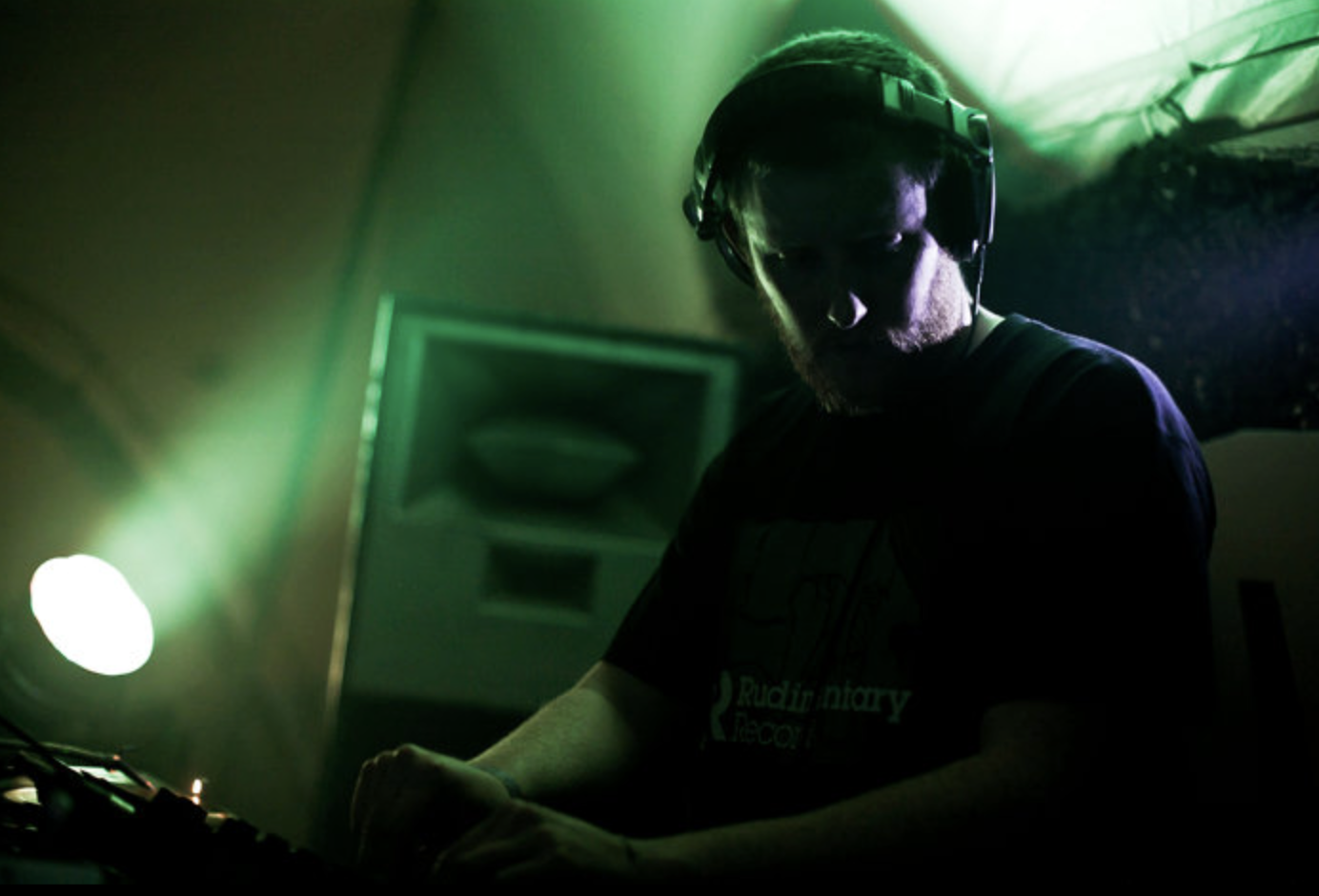 A DJ wearing headphones is working at a DJ booth in a dimly lit room with green lighting.