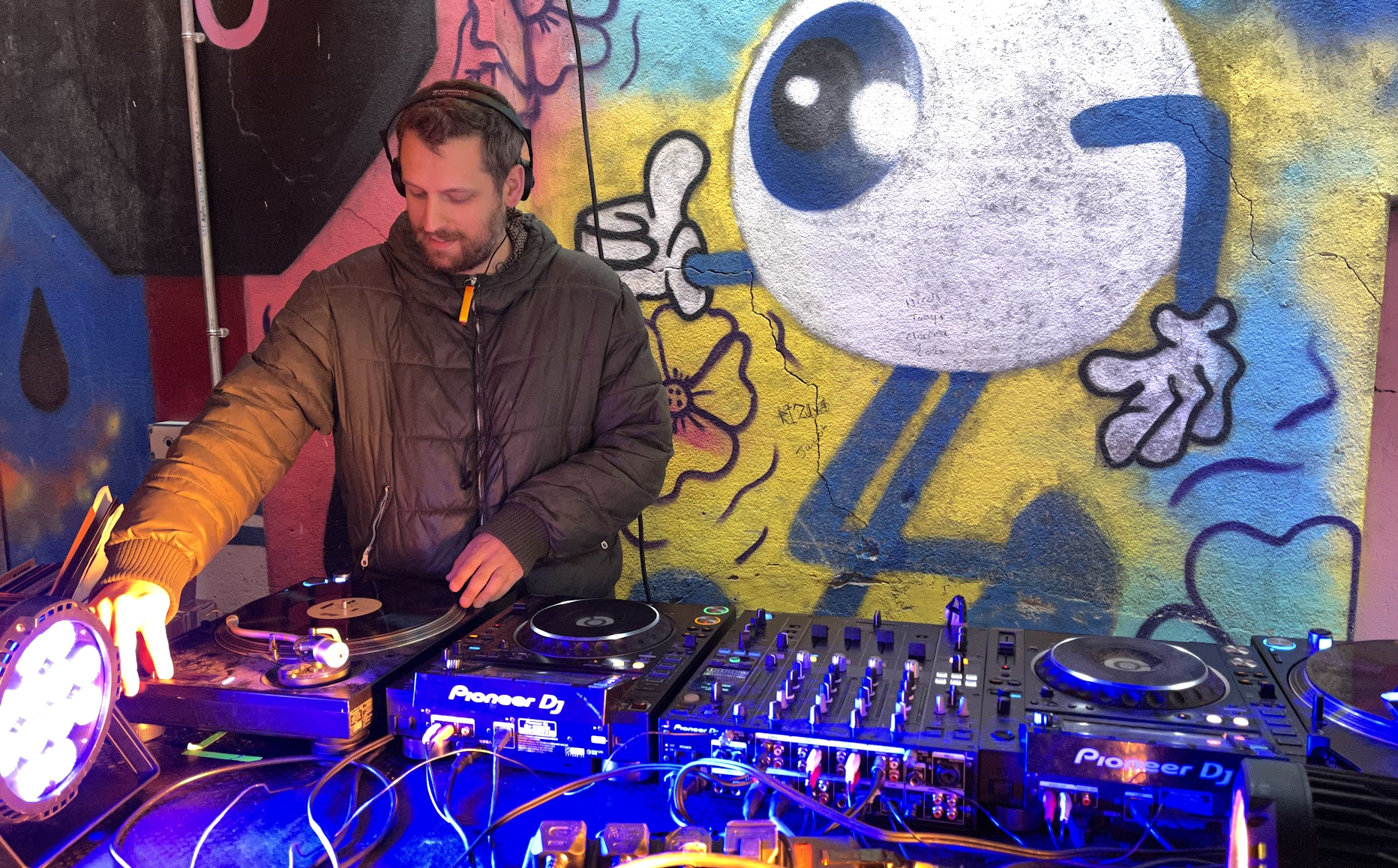 A man wearing a black jacket and headphones is DJing at a club with colorful graffiti art, including a large cartoon face and various shapes, painted on the wall behind him.