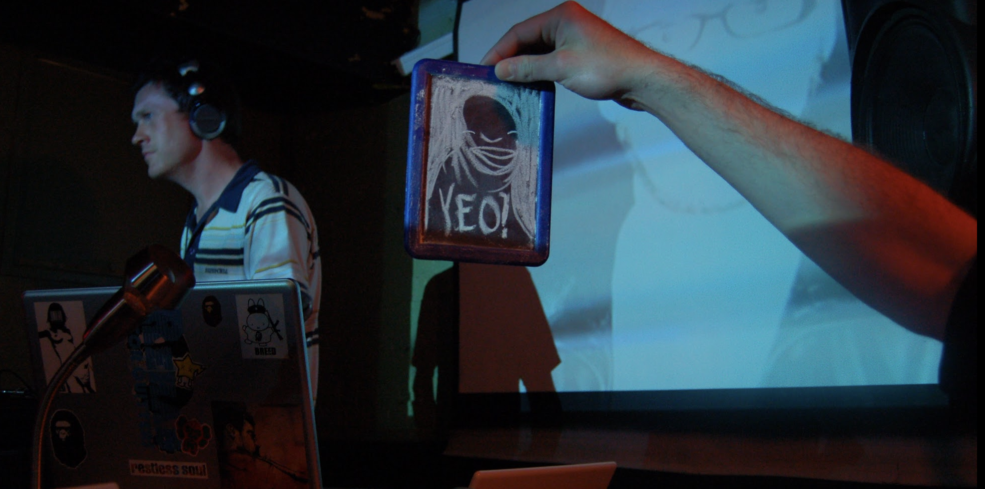 A DJ wearing headphones is in front of a laptop decorated with stickers. A person's hand is holding a small rectangular object with drawn art and the word 'YEOH' written on it in front of a projection screen.