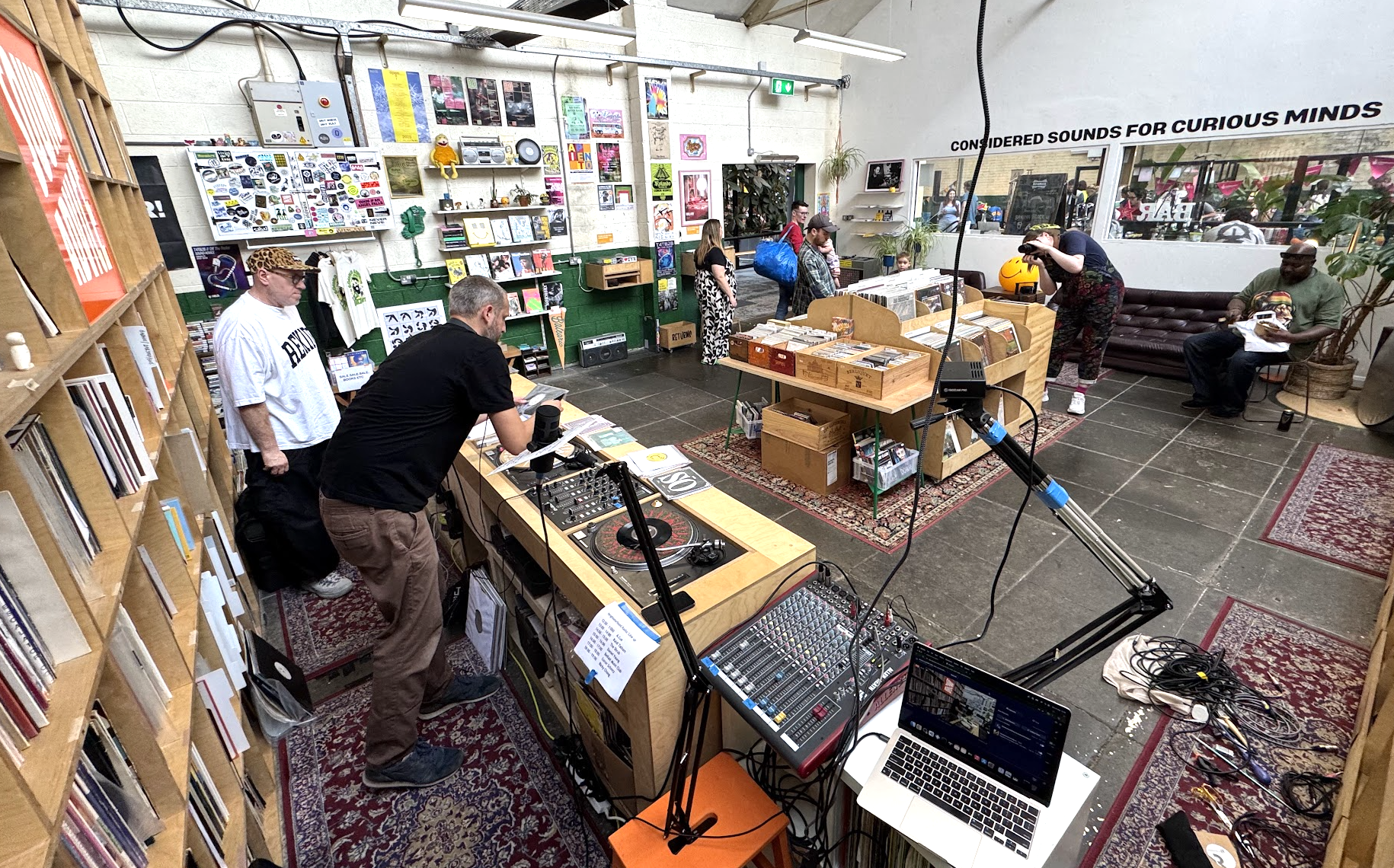 A busy record store with vinyl records, books, and posters on the walls. People browse, take photos, and listen to music. DJ equipment, a laptop, and audio gear are set up at the front.