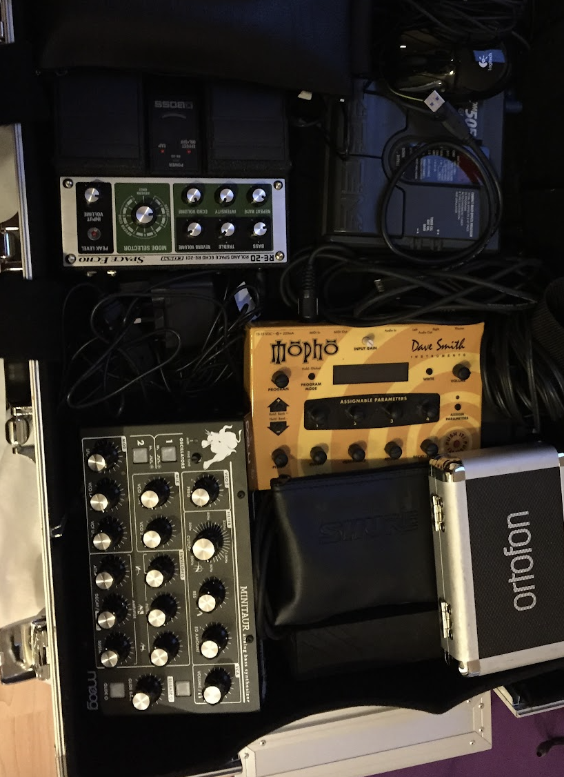 A collection of music equipment including a Moog Minitaur synthesizer, an M-Audio MIDI controller, a Dave Smith Instruments Mopho synthesizer, an S2 Audio guitar amplifier, a Boss effects pedal, and various cables and a black leather pouch.