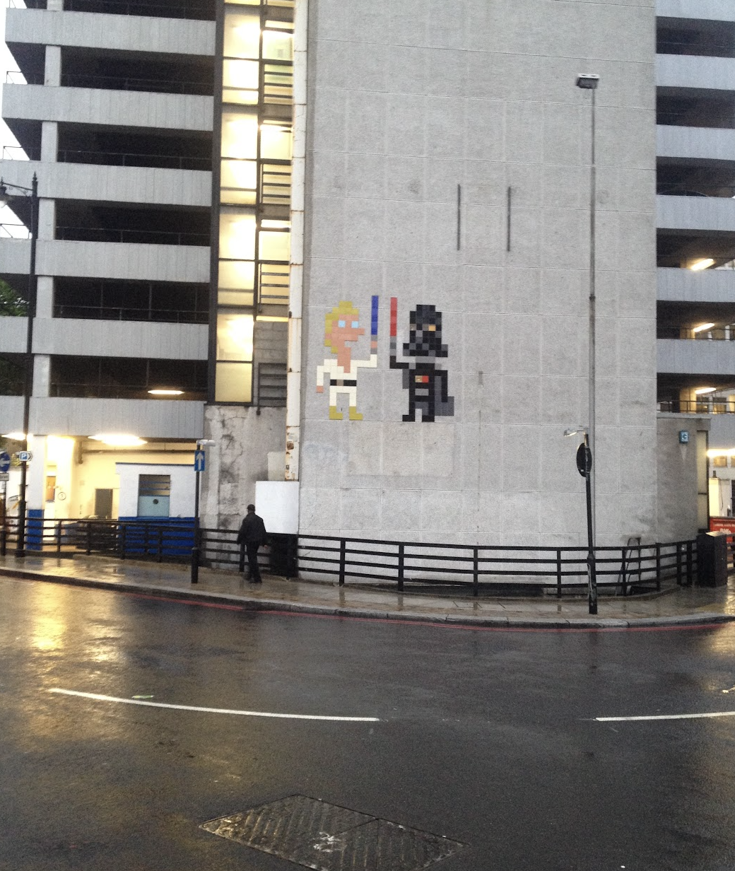 Pixel art mural of a blonde woman in a white outfit holding a red and blue lightsaber and a black figure with a cape, also holding a lightsaber, on a gray building wall.