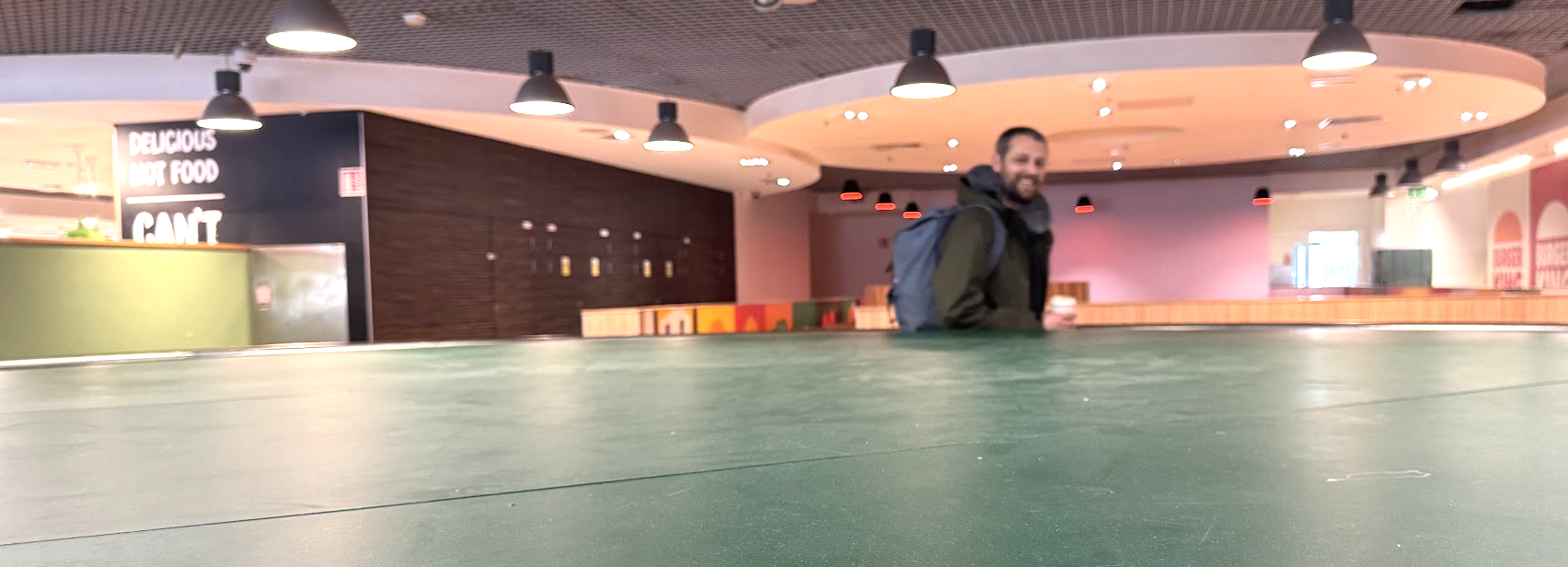 A man with a backpack smiling while standing inside a modern indoor space, possibly a shopping mall or airport terminal, with ceiling lights and colorful decor in the background.