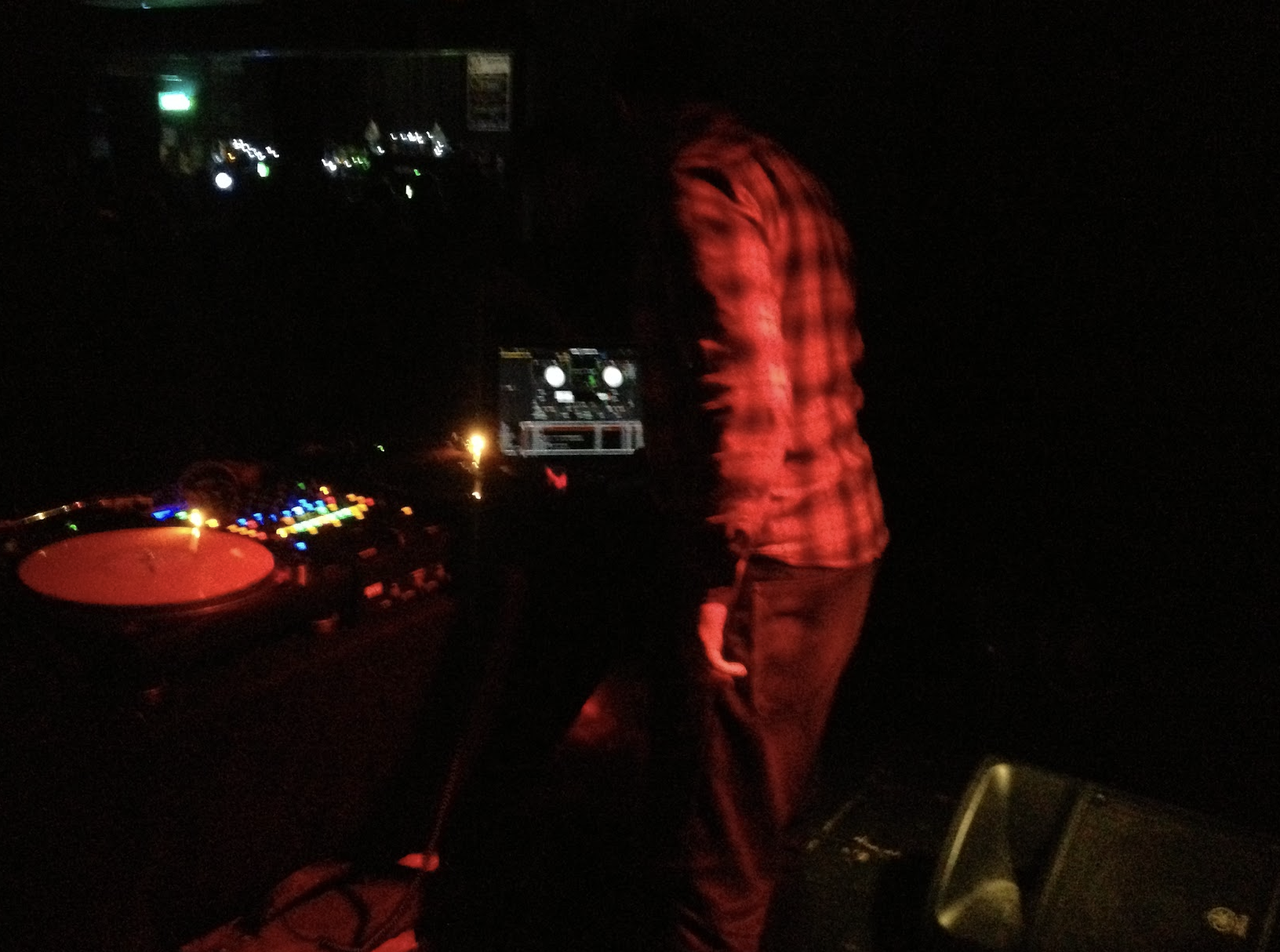 A DJ wearing a red plaid shirt is performing at a dark nightclub or event, with DJ equipment and colorful LED lights on the table.
