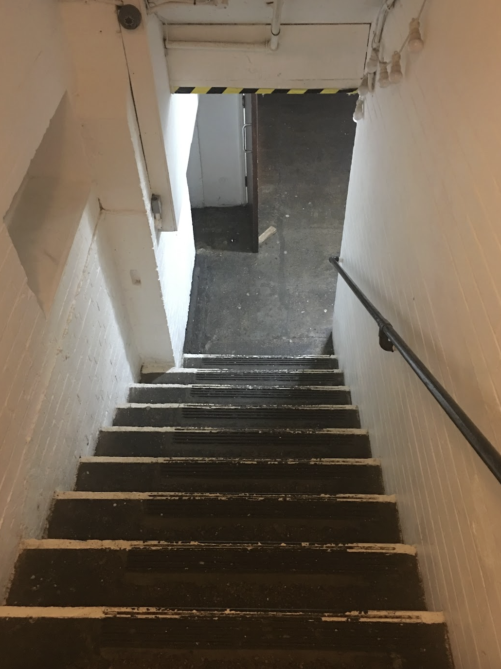Interior stairwell with black steps, white walls, metal handrail, and an open door at the top leading to a dark corridor.