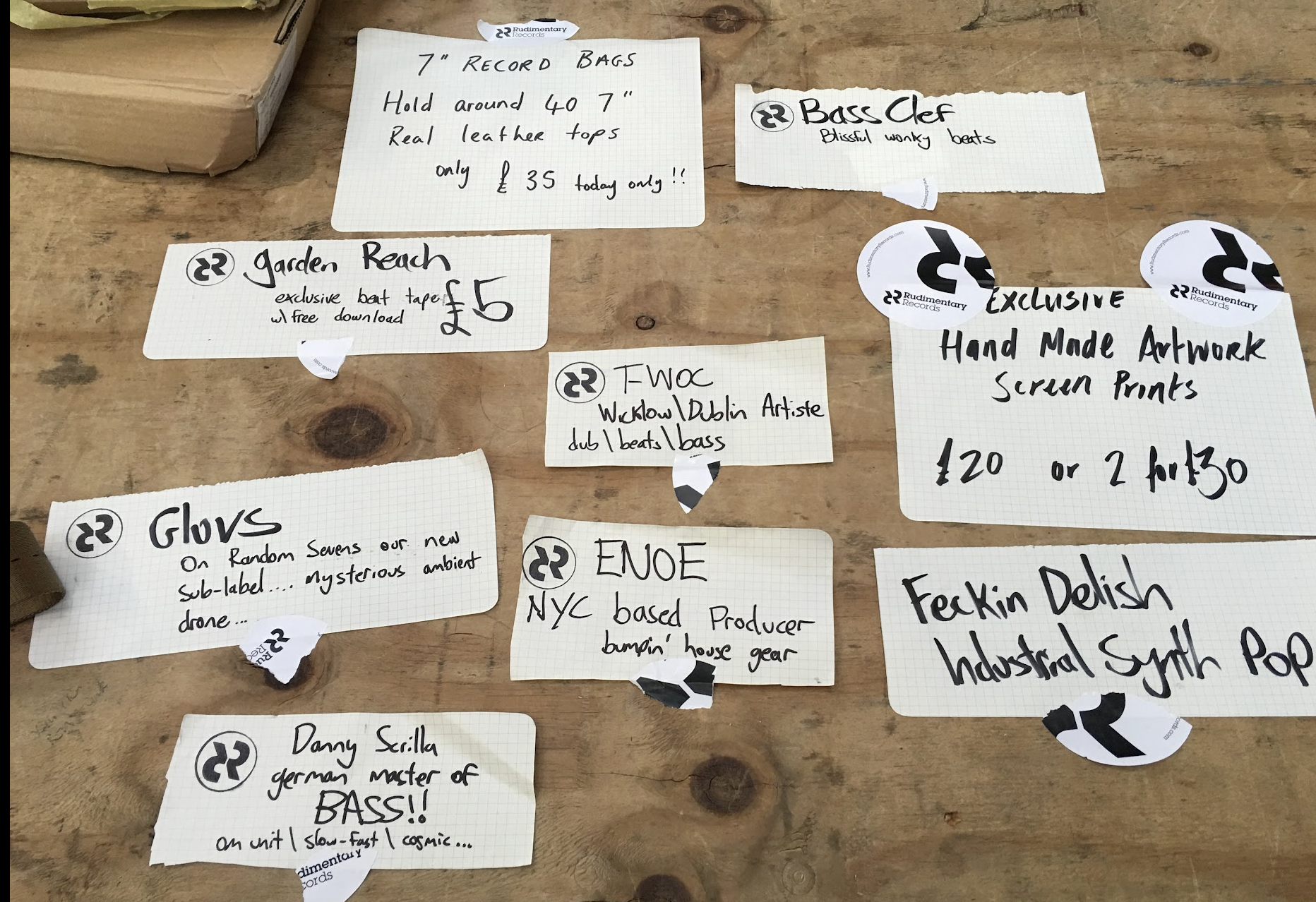 Several handwritten notes on torn graph paper with various product descriptions and prices, scattered on a wooden surface. Some notes are from Rudimentary Records, indicating music-related items and artwork.