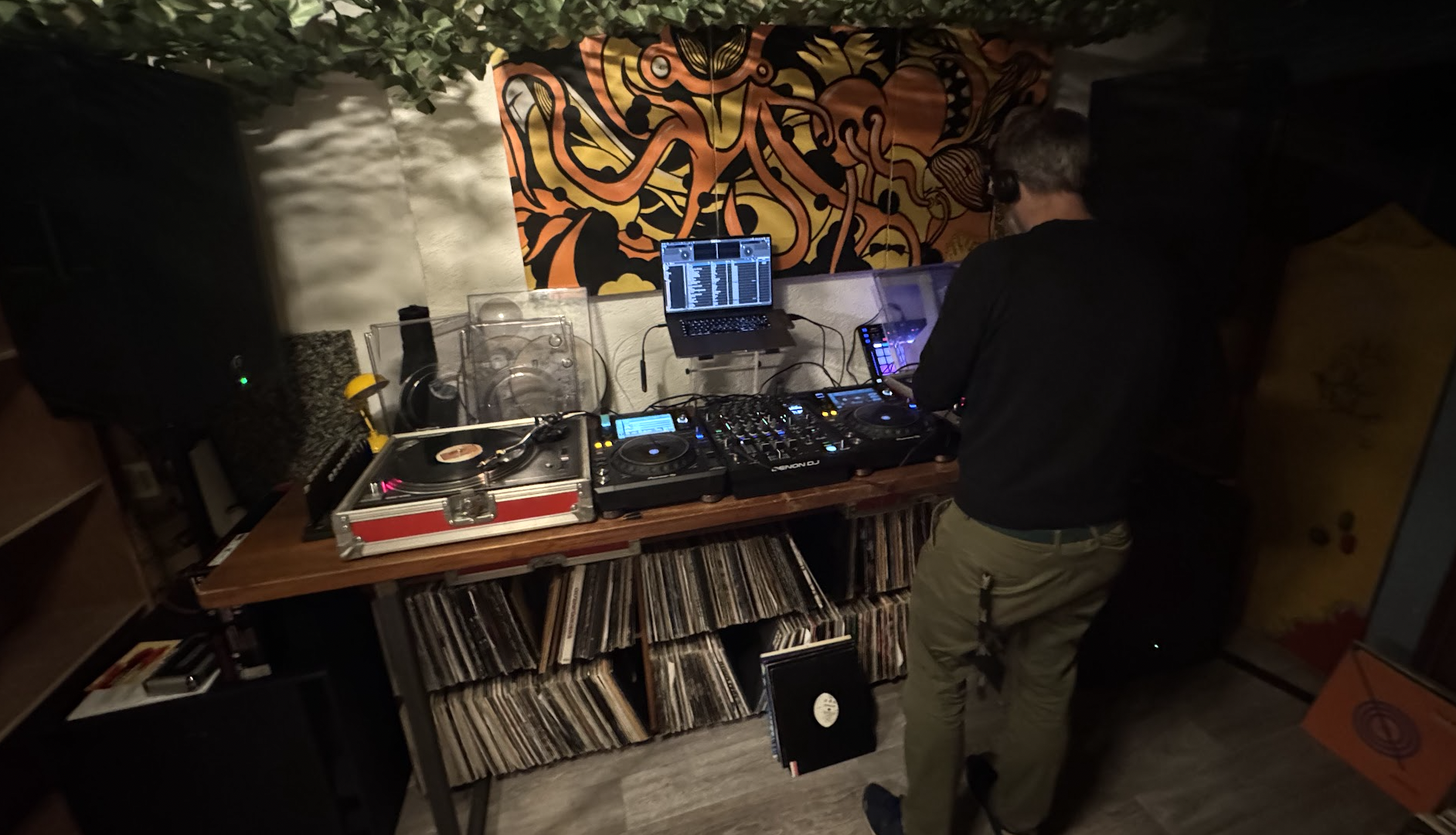 A person DJing with turntables, a mixer, and a laptop in a room decorated with a colorful abstract wall hanging.
