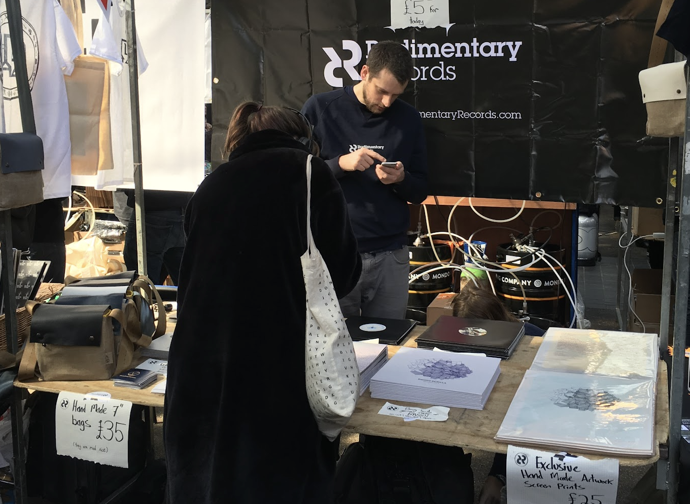 A street market stall selling handmade bags and exclusive hand-made artwork screen prints. Two people are browsing the items, one person is using a phone, and there are signs with prices for the items.