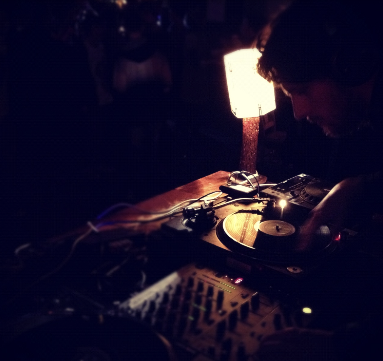 A DJ performs in a dimly lit room, using turntables and a mixer, with a small lamp providing warm light.