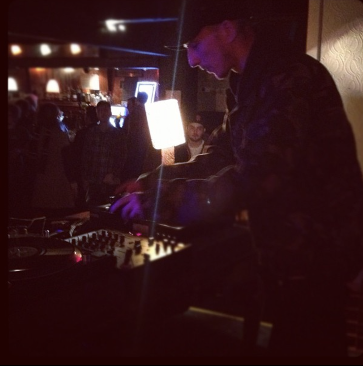 A DJ plays music in a dimly lit bar or club with people in the background, some sitting and some standing, under colorful lights.