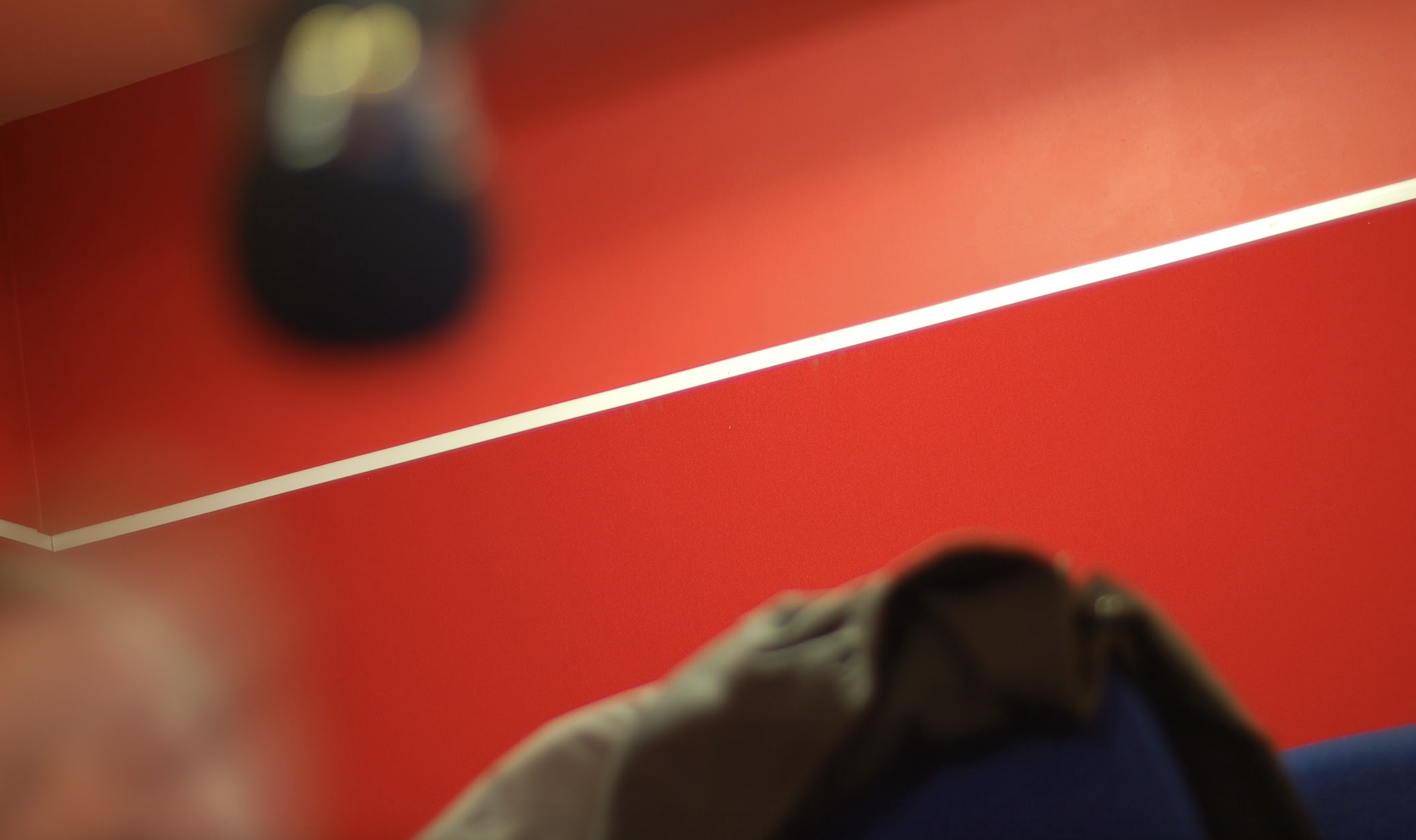Close-up of a red table with a white line, a dark object blurred in the background, and part of a person's shoulder and clothing in the foreground.