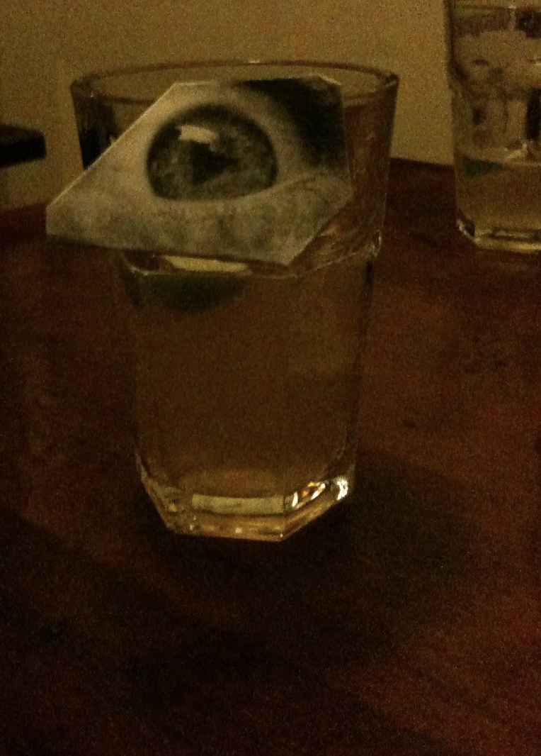 A glass of whiskey with a dollar bill folded and placed on top of it, on a wooden table.