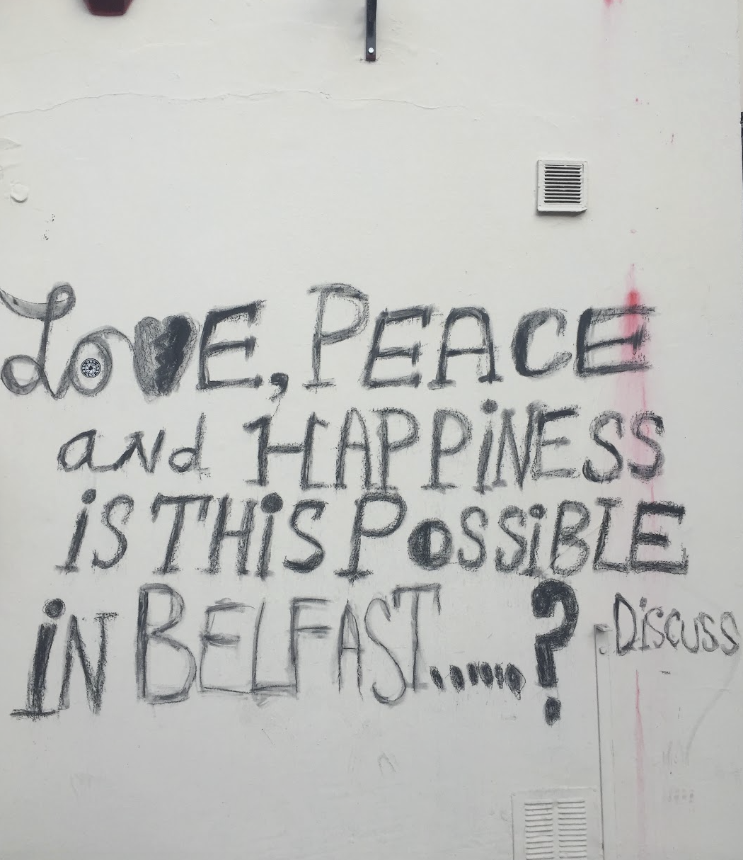 A wall with a handwritten message in black spray paint that reads: "Do love, peace and happiness is this possible in Belfast?"