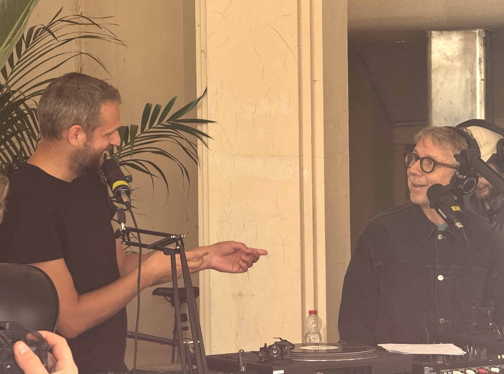 Two men in a recording studio, one with gray hair and glasses, speaking into a microphone, and the other with gray hair and a beard, listening and smiling, surrounded by professional audio equipment and a plant in the background.