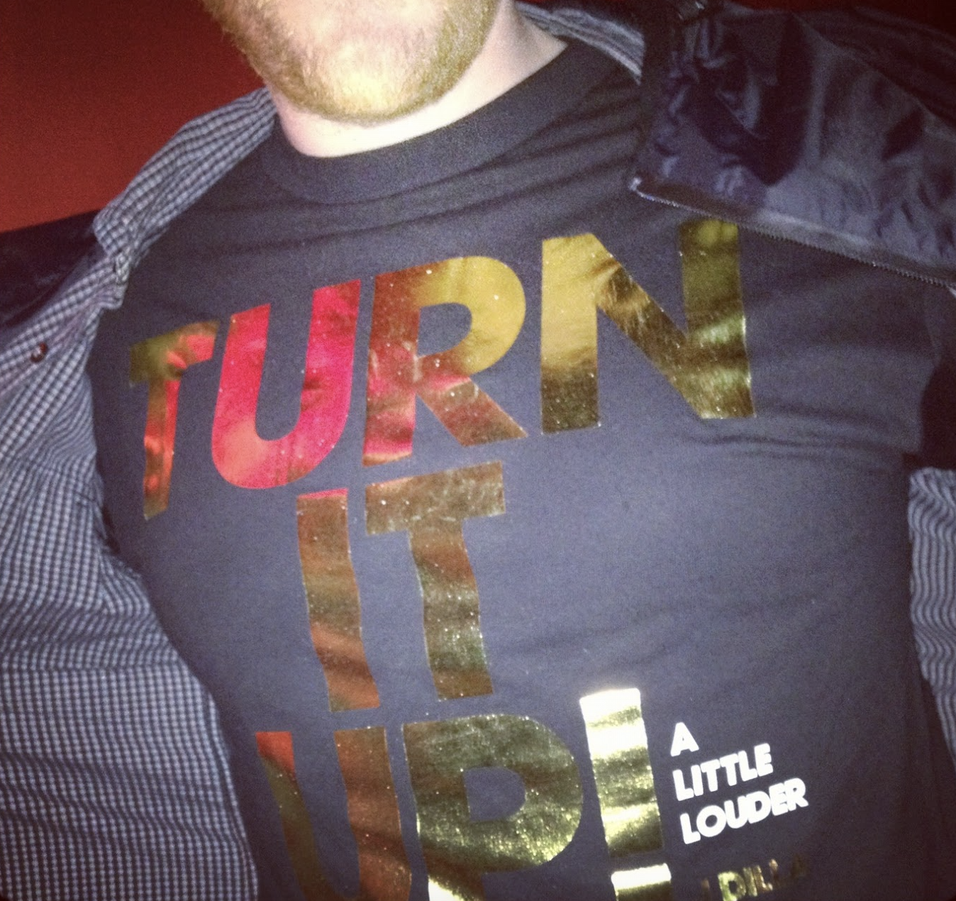 Close-up of a person wearing a black T-shirt with shiny gold and red lettering that says 'TURN IT UP! A LITTLE LOUDER', with some of the person's face and arms partially visible.