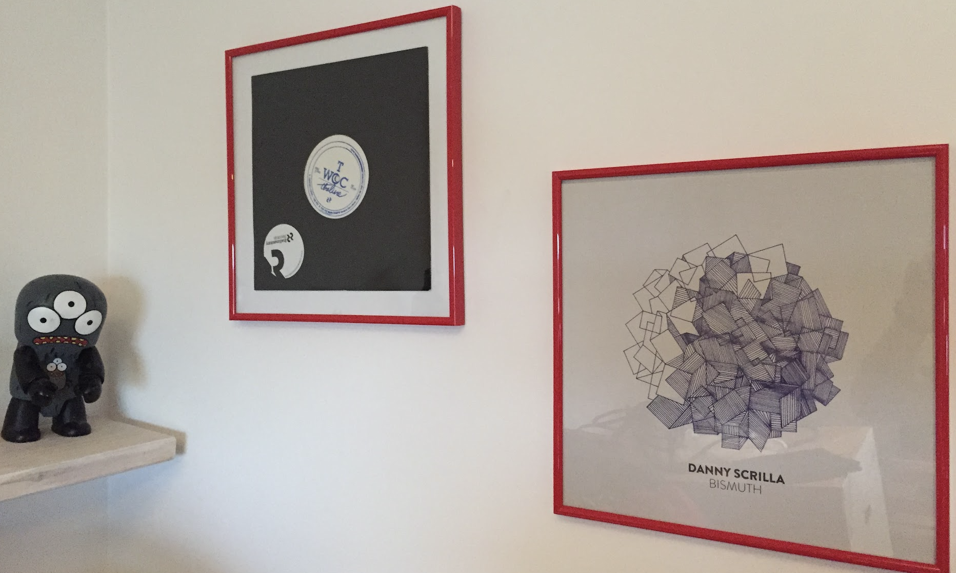 Decorative wall art and a toy figure. The wall art includes a framed black and white circular design with text, and a framed abstract geometric drawing with the caption "DANNY SCRILLA BIMUTH." There is also a plush toy figure with three eyes, an open