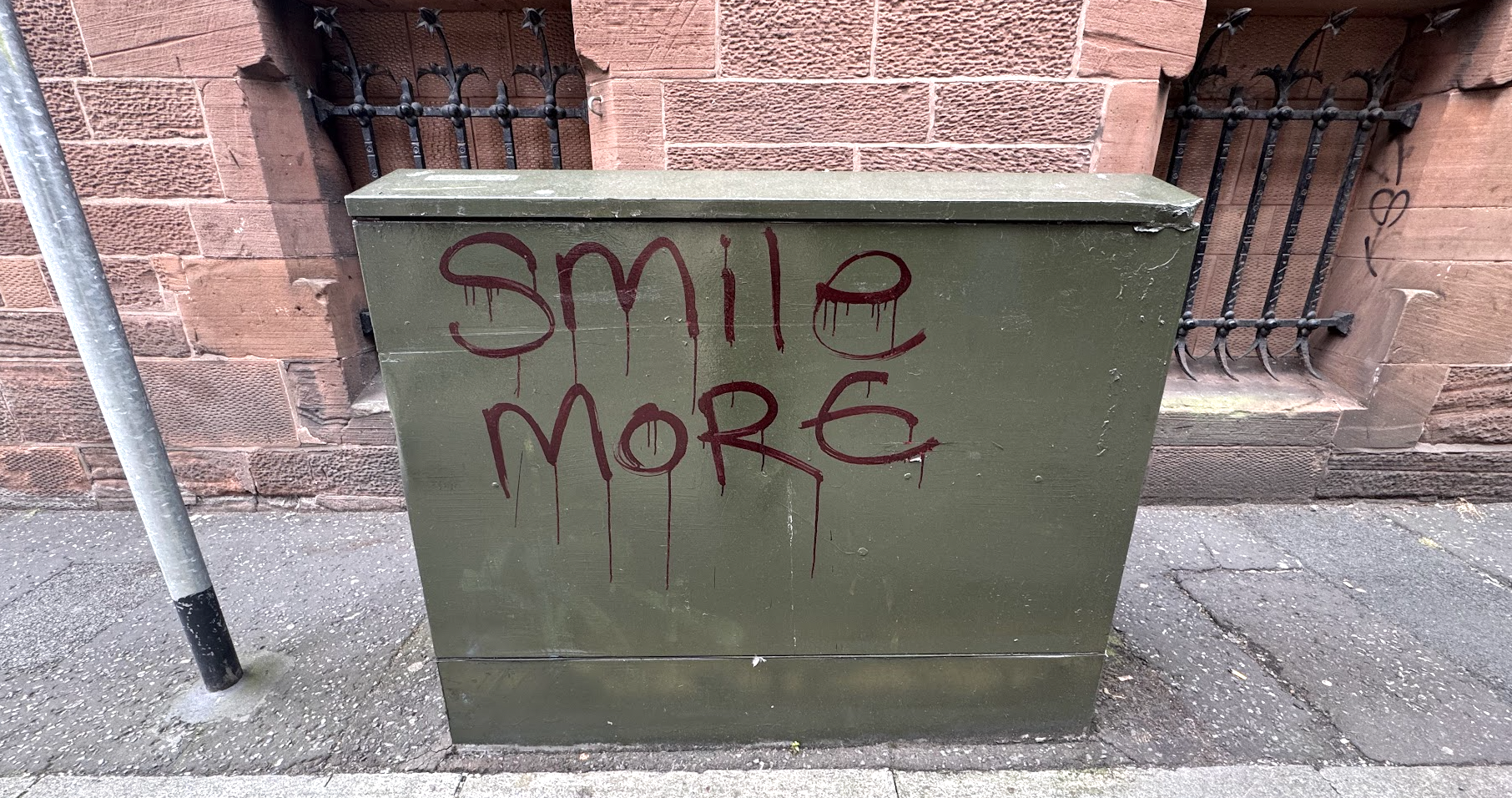 Graffiti on a green utility box that reads 'smile more' in red spray paint, situated on a sidewalk against a red brick wall with black iron bars.