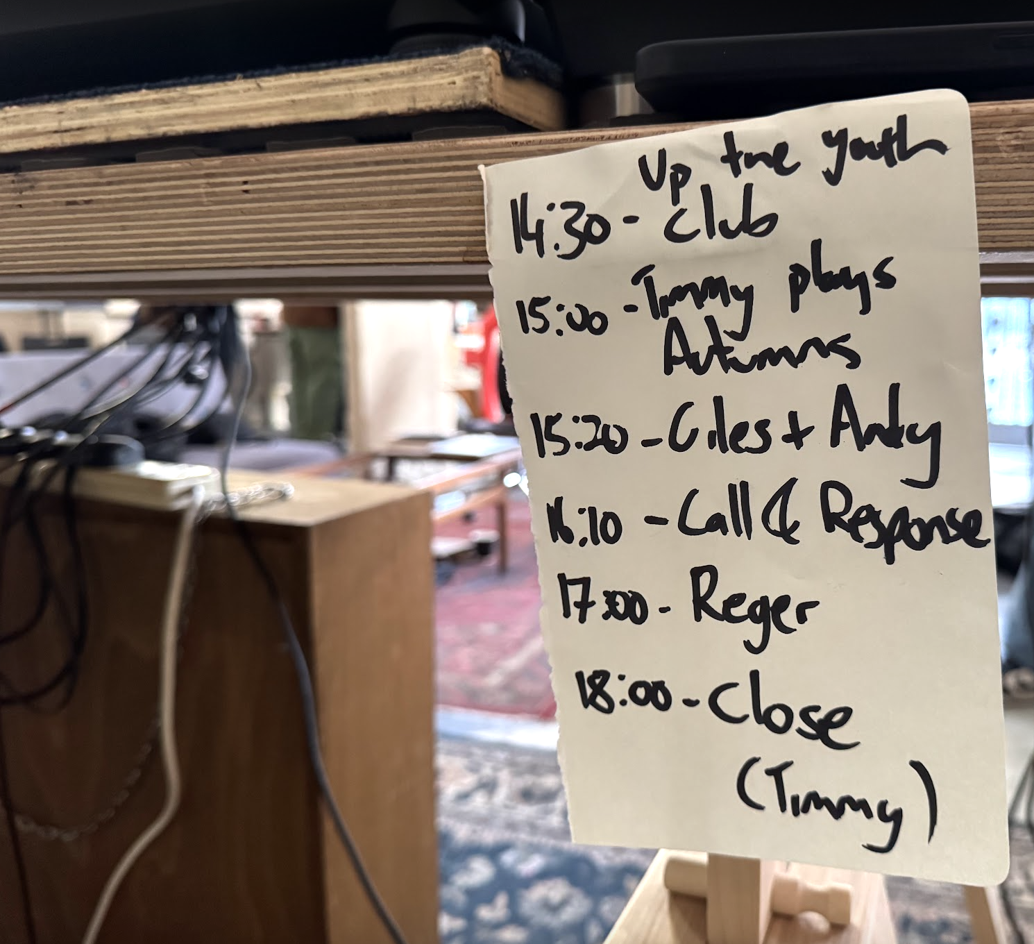 A handwritten schedule on a sticky note listing various activities and times, attached to a wooden structure in a casual indoor setting with blurred furniture and a patterned carpet in the background.