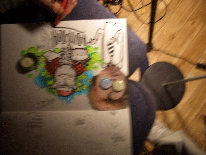Close-up of a hand holding a coloring book with a drawing of a cartoon character and three small containers of what appears to be paint or stickers, on a table with a wooden floor in the background.