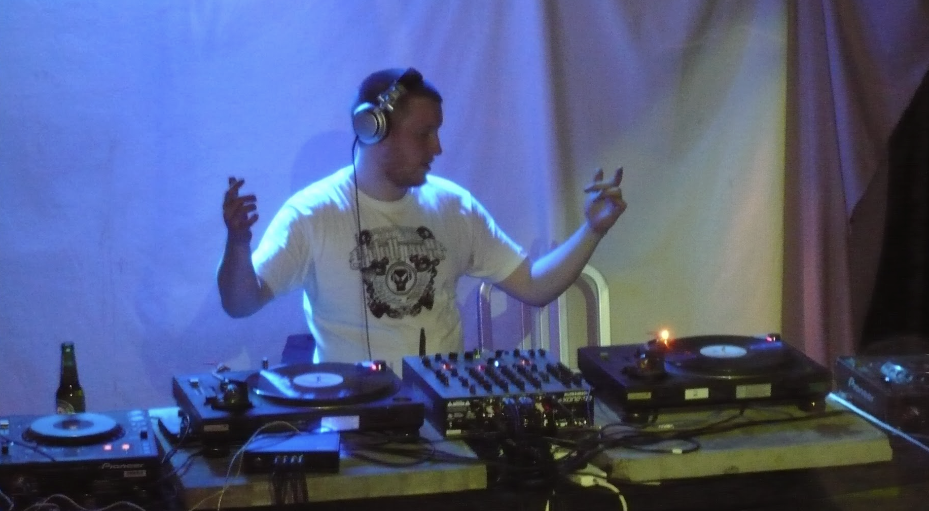DJ wearing headphones, playing music at a DJ booth with turntables and equipment, with a bottle on the table. The background has colorful lighting.
