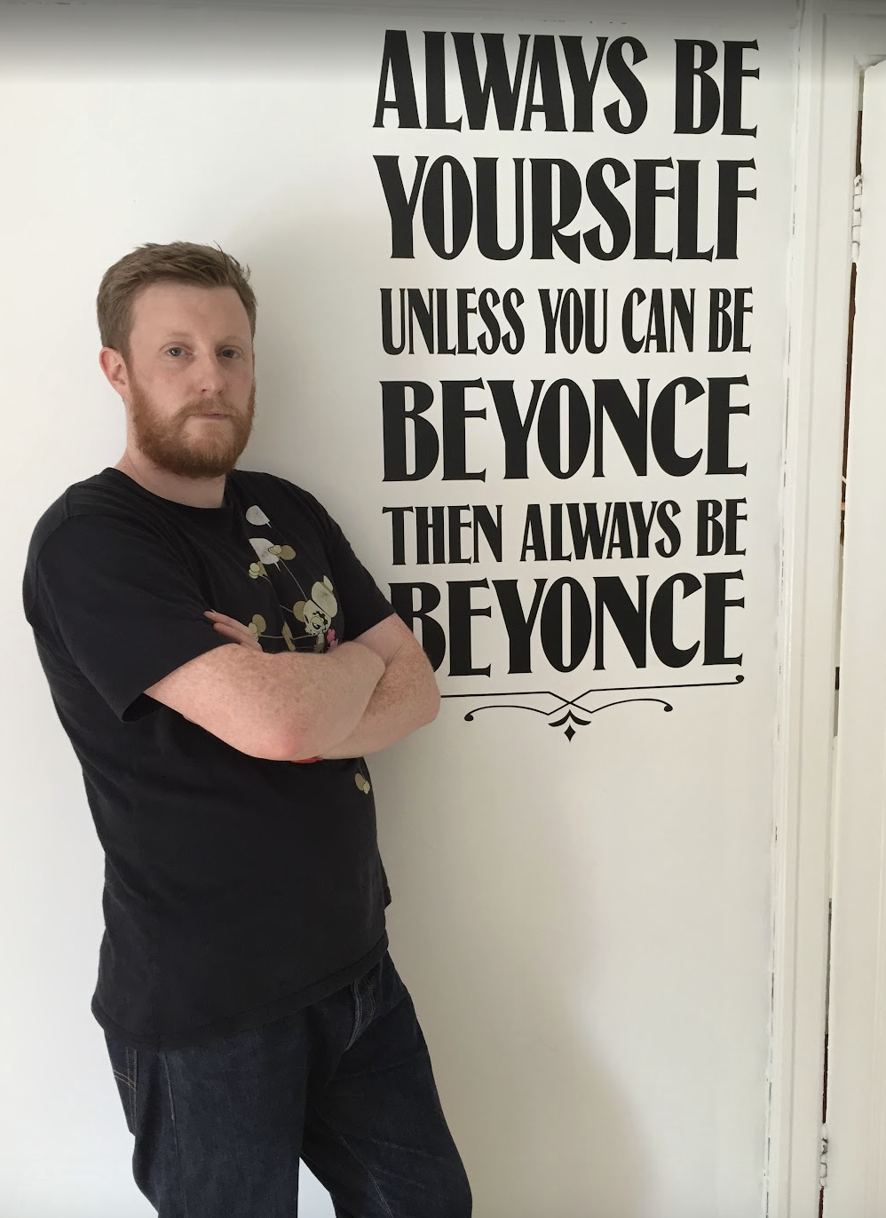 A man with red hair and a beard stands with arms crossed next to a wall with a large quote about being yourself and Beyoncé.