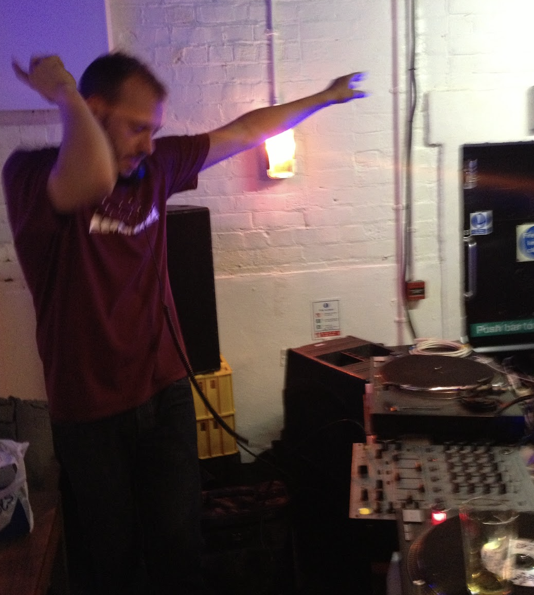 A man with headphones dancing behind DJ equipment in a dimly lit room with purple and pink lighting.