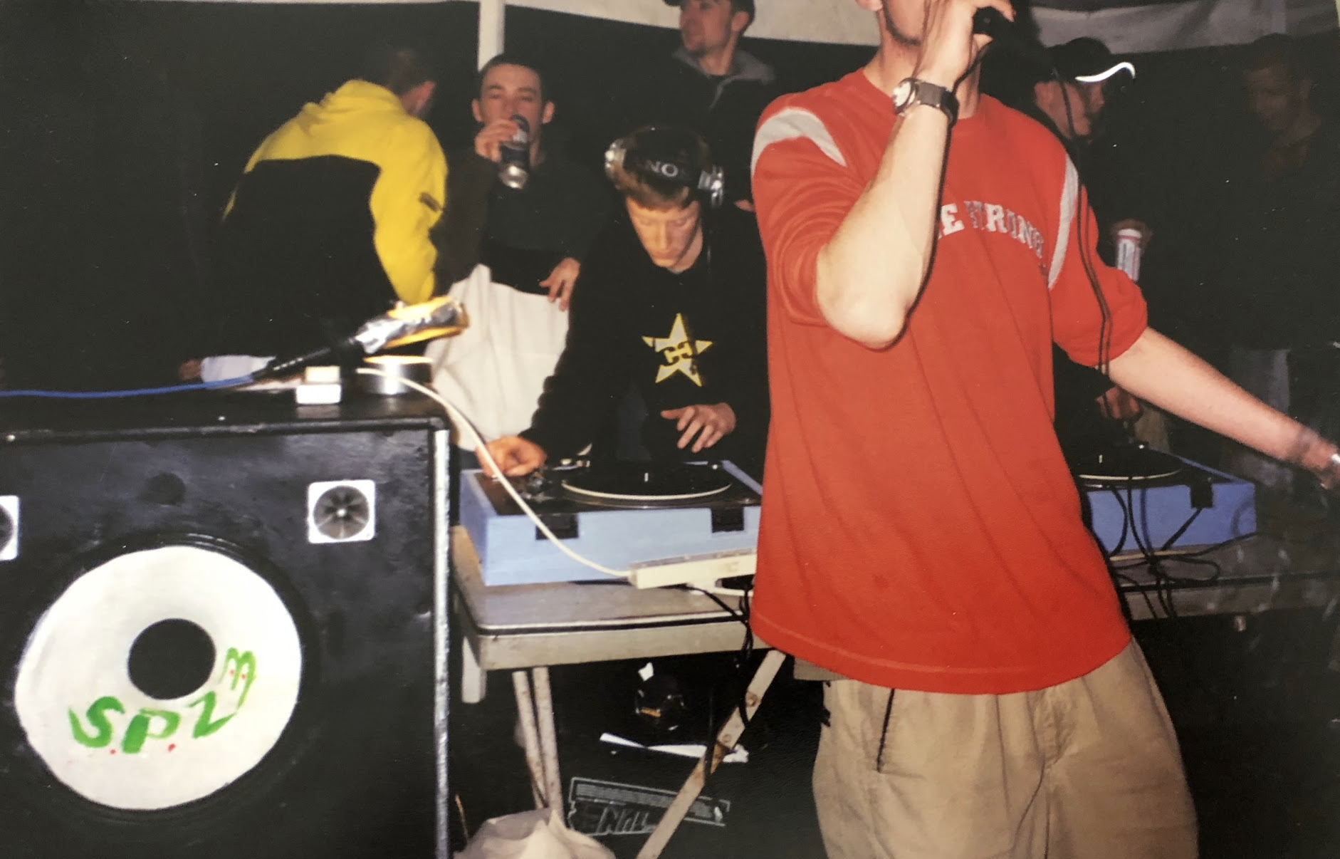 Group of people with DJ equipment and large speaker at a party or dance event, some wearing headphones and casual clothes.