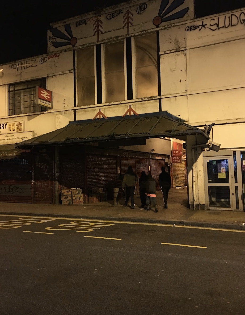 Night street scene in an urban area with closed shops and pedestrians walking past. A building with graffiti and a sign indicating a subway station. Dim lighting illuminating the scene.