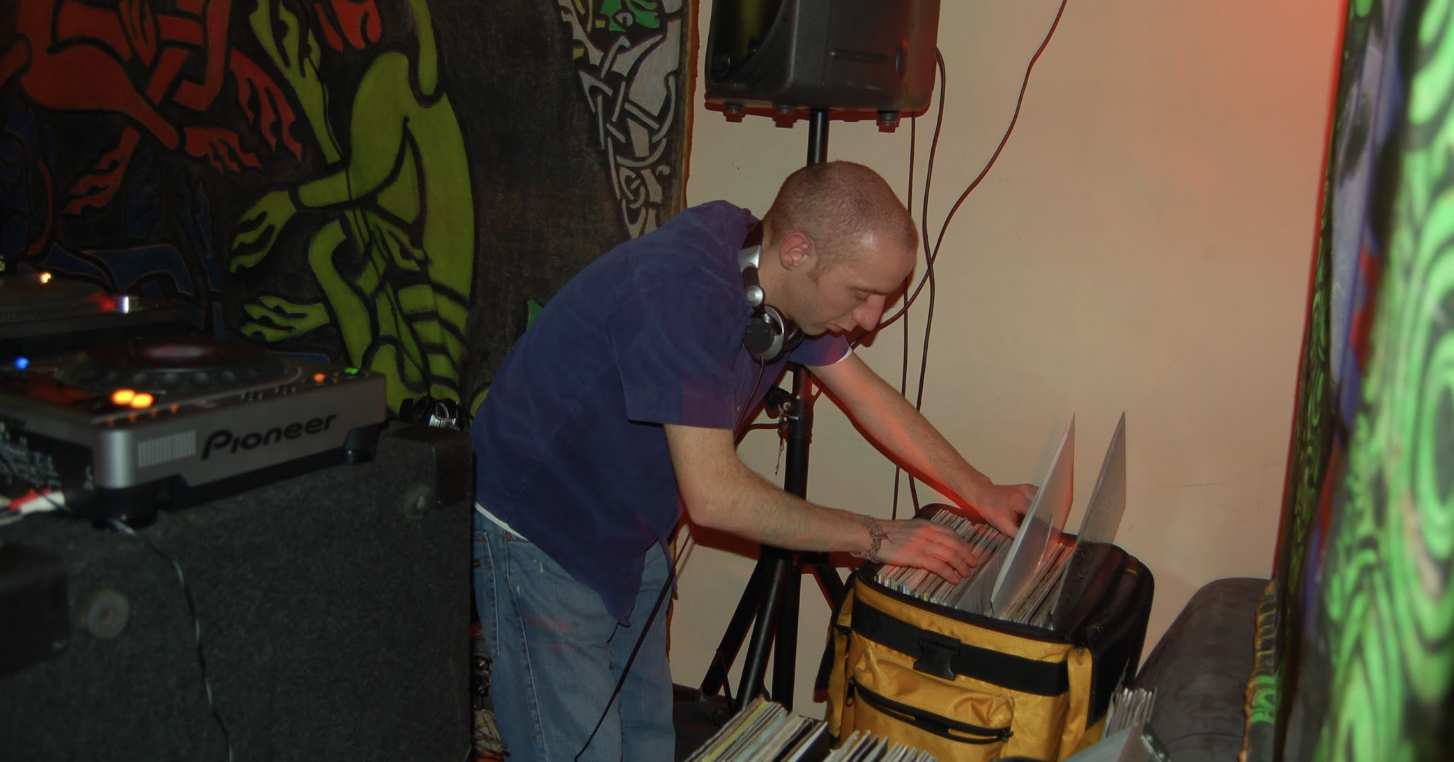 A DJ with a shaved head wearing headphones, a dark blue shirt, and jeans, is organizing vinyl records from a yellow bag. The scene is set in a room with colorful graffiti art on the walls, and DJ equipment, including a Pioneer turntable and a speaker
