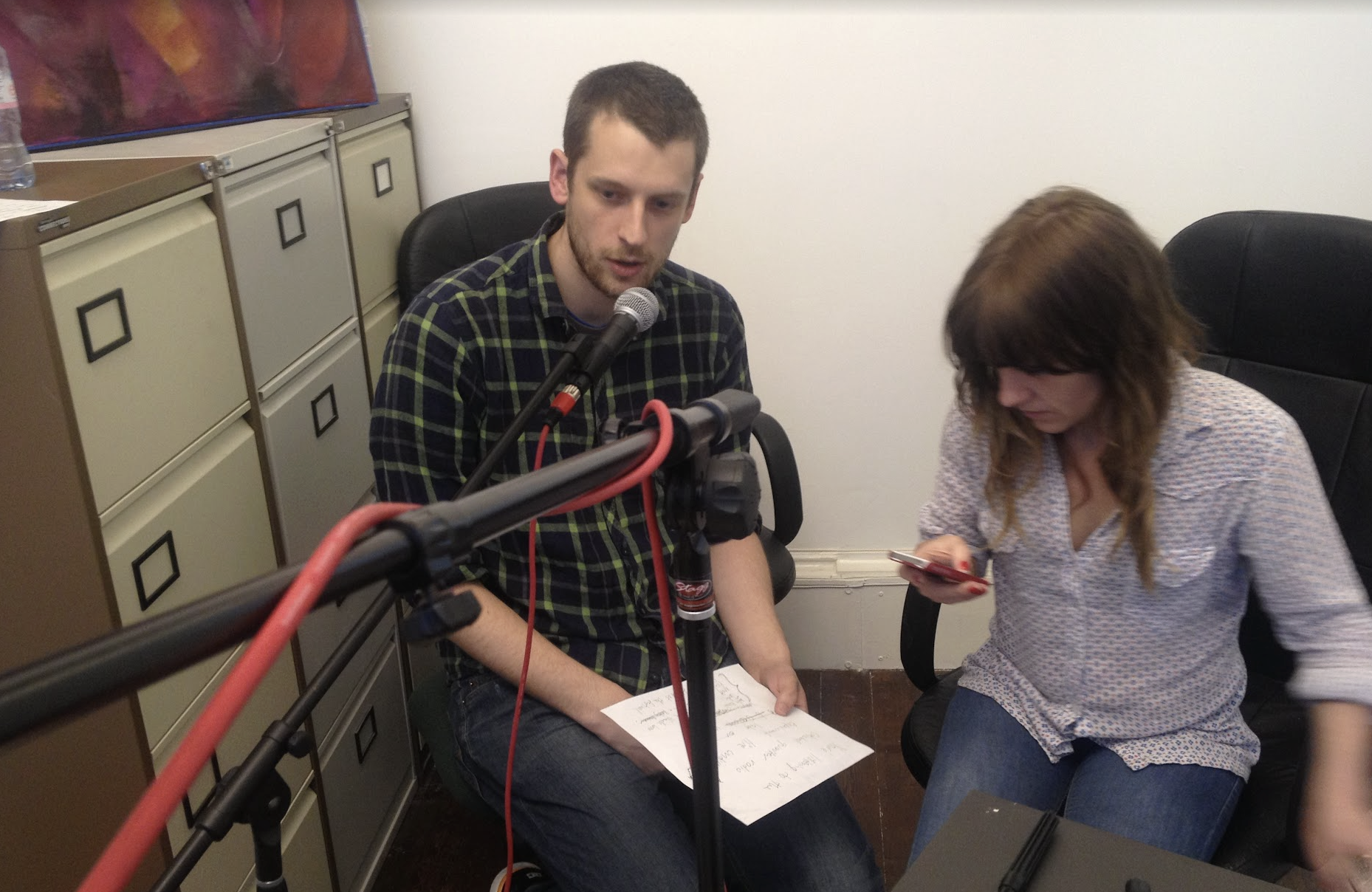A man and a woman are in a room with a microphone on a stand and a camera pointing towards them for recording. The man has short brown hair, a beard, and is wearing a black and green checkered shirt. He is sitting in a black office chair and holding