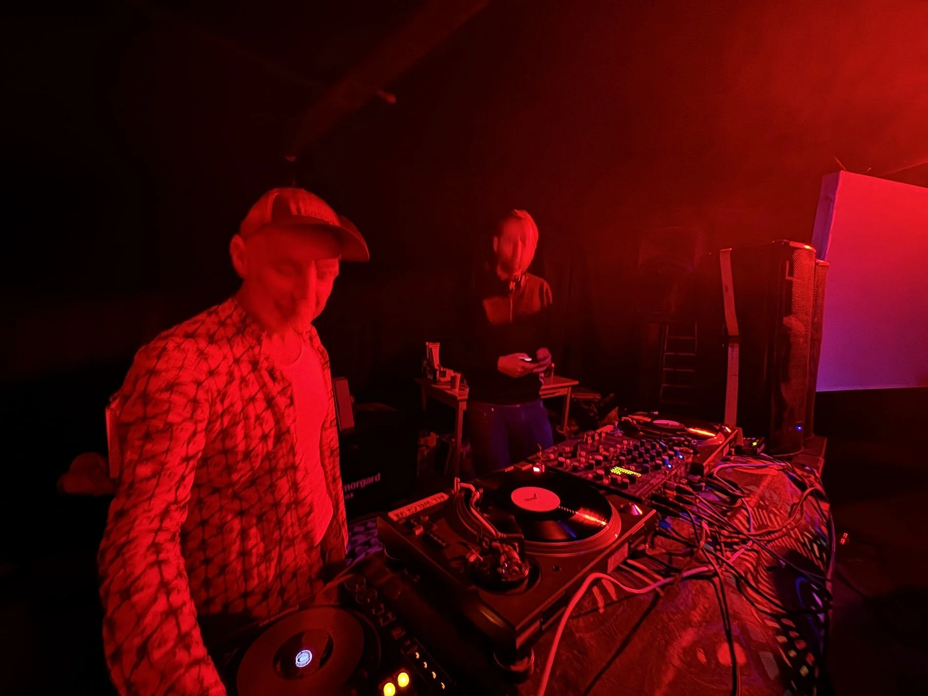 Chuck and Andy at a DJ booth with turntables and equipment, at a warehouse rave.
