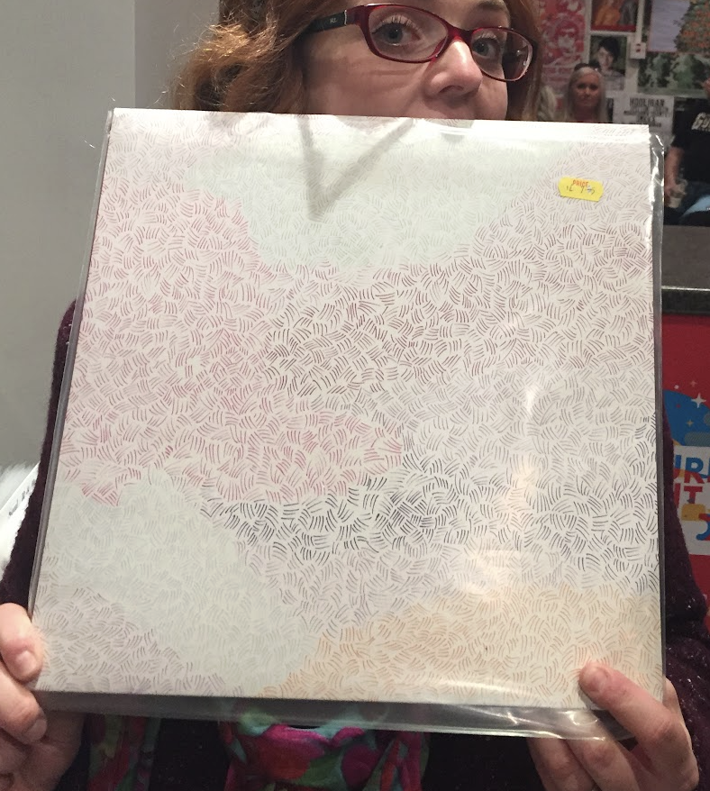 Person with glasses holding a large framed print with a pattern of small, colorful, overlapping leaves or feathers. A yellow price sticker is visible on the top right corner of the frame.