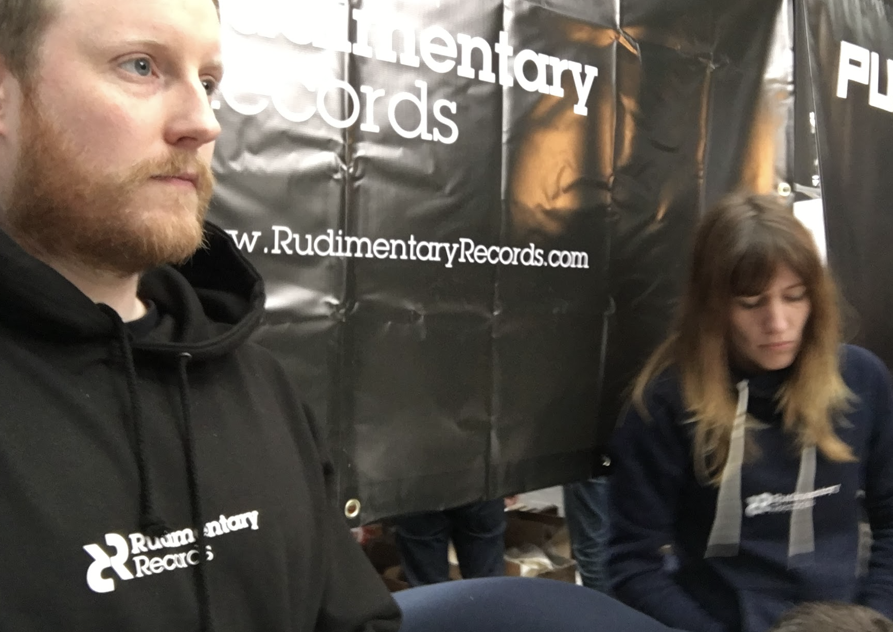 Two people sit in front of a black banner with white text that reads 'Rudimentary Records' and a URL 'W.RudimentaryRecords.com'. The man on the left has a light beard, blonde hair, and is wearing a black hoodie with the same logo. The woman on the ri