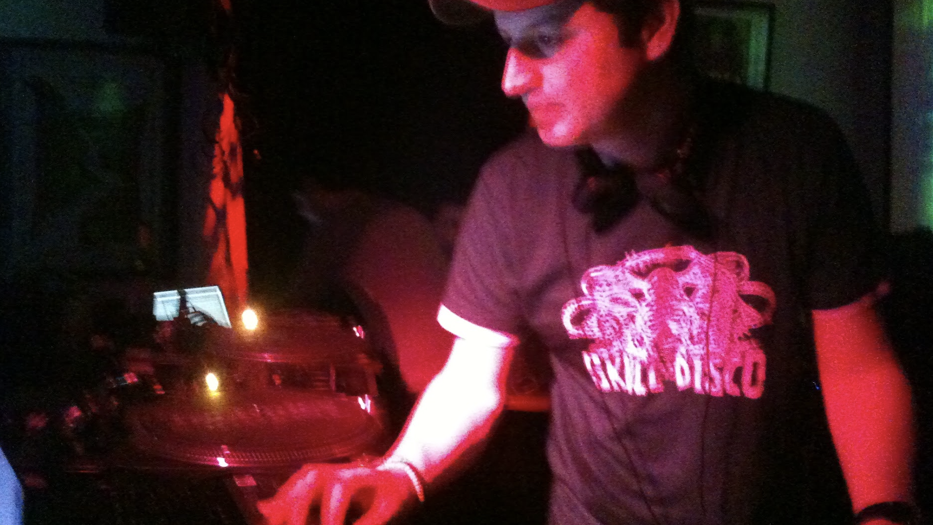 A DJ wearing a black T-shirt with a graphic and the words 'DANCE DISCO' is working with DJ equipment in a dark setting with red and green lighting.