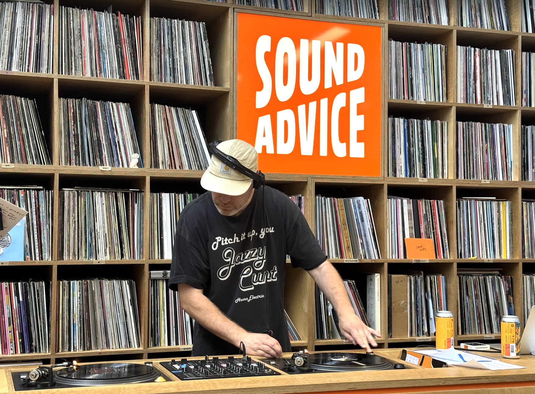 A DJ wearing a black t-shirt and a beige cap, with headphones, is playing music on a turntable in front of a wall of vinyl records. A large orange sign with white text reads 'SOUND ADVICE' behind him.