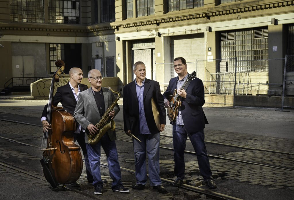 Upright bass player, saxophonist, drummer holding sticks and a cymbal, and guitarist on train tracks in a downtown.
