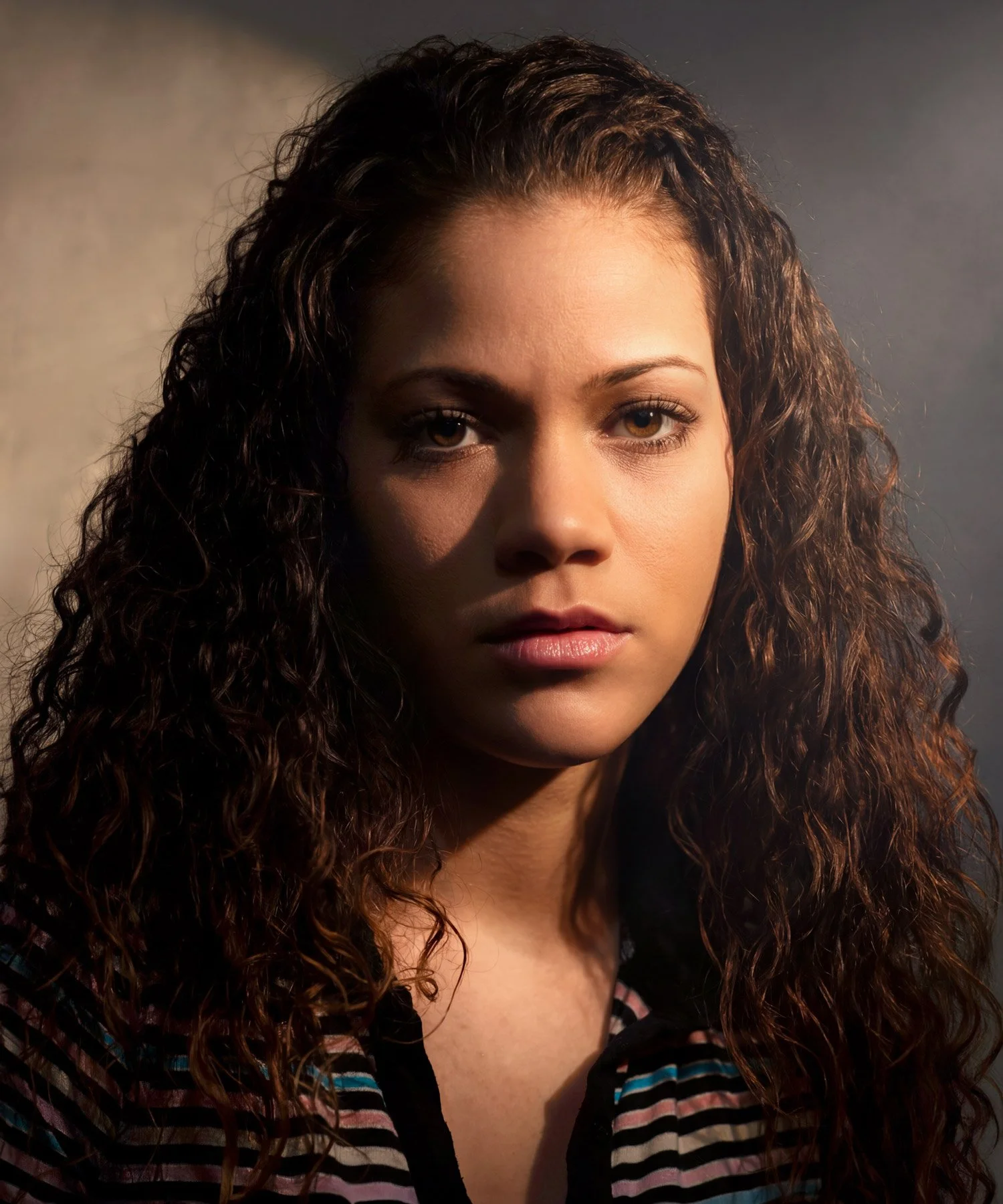 A young Latina female with long, curly hair. and blue, pink, white, and black striped shirt gazes intently into the camera. Shot with directional, high-contrast cinematic light.